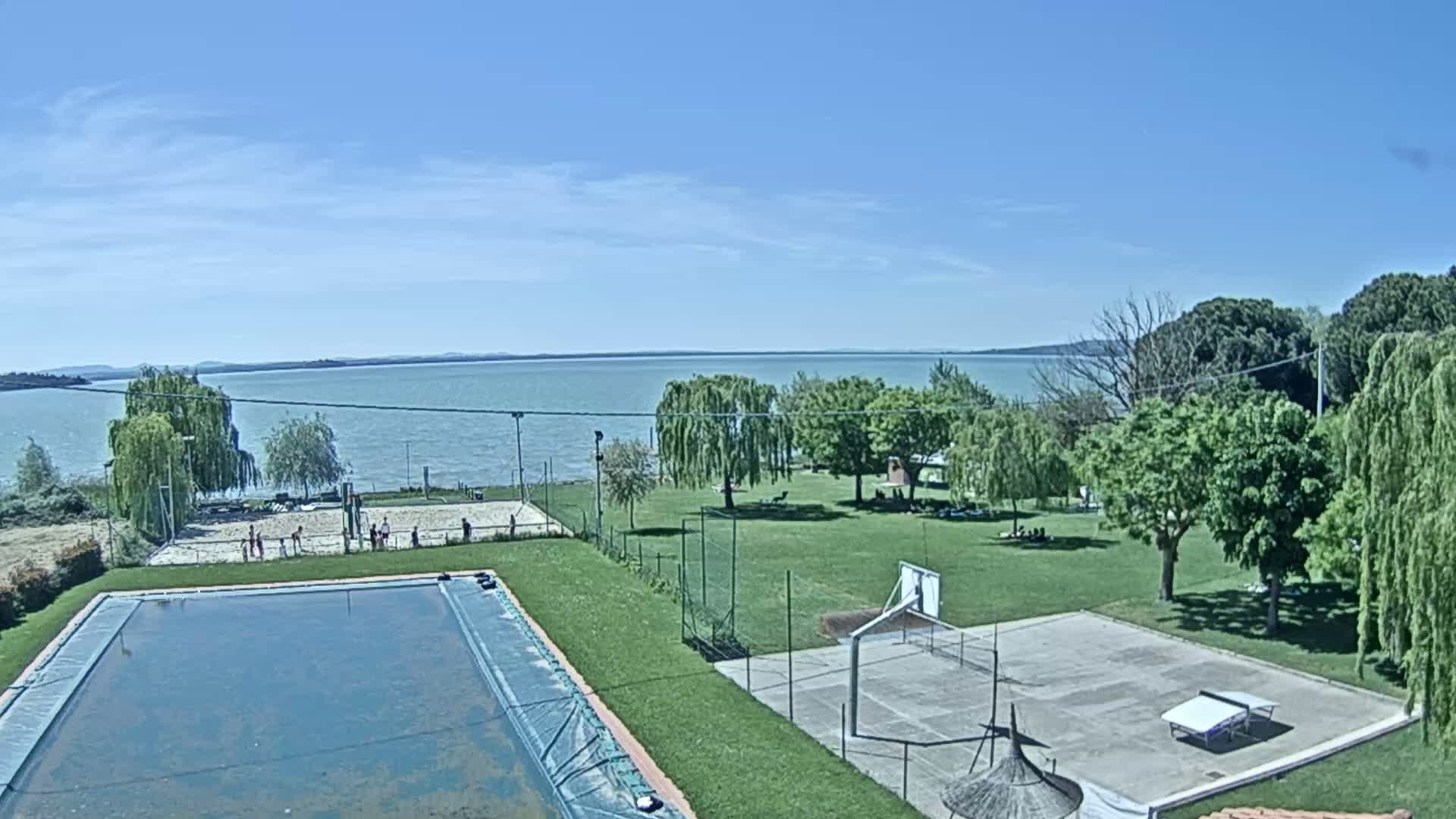 A swimming pool and outdoor courts for volleyball, basketball, and table tennis are situated on a grassy area overlooking a calm lake under a clear blue sky.