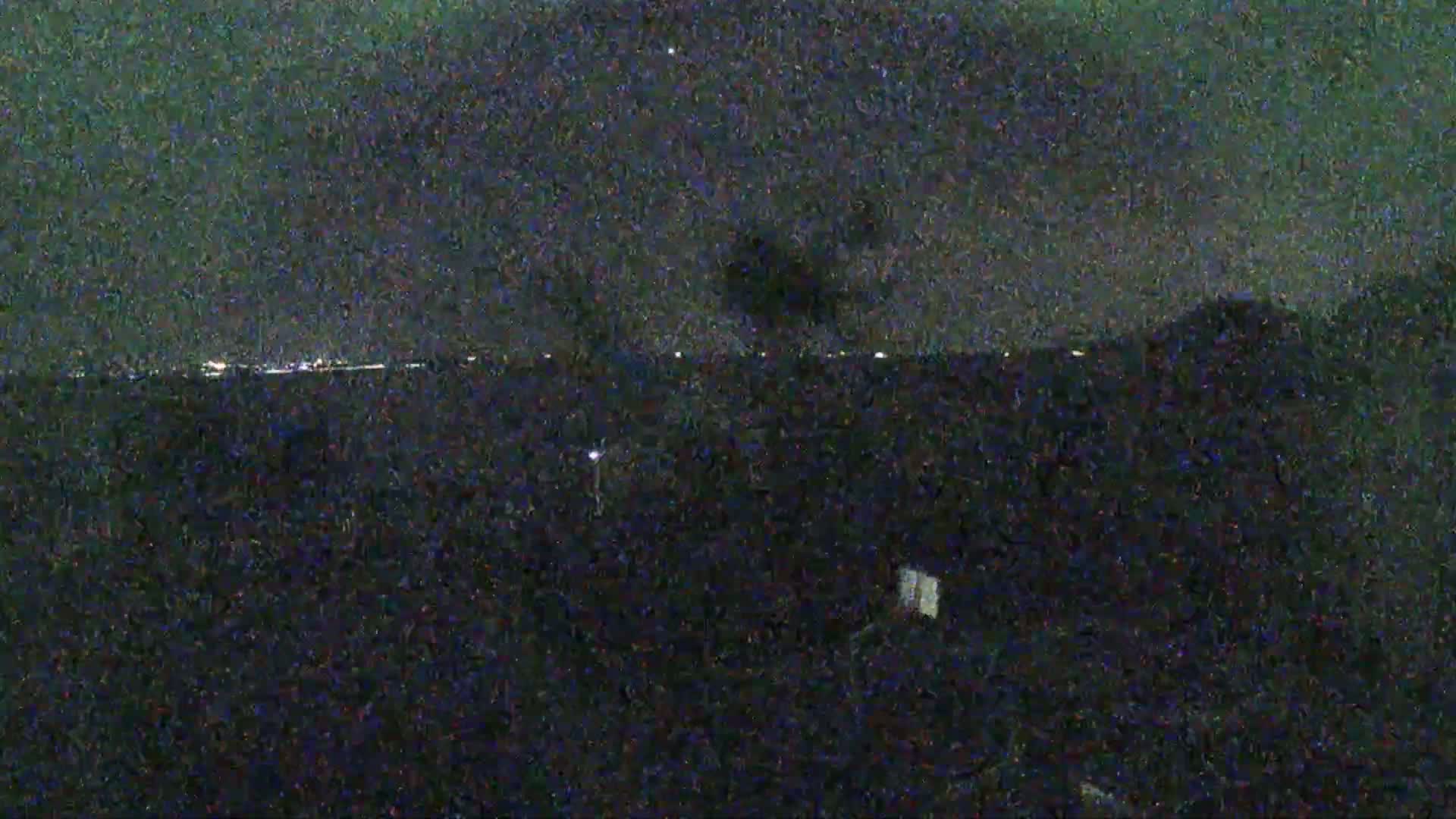 A nighttime view shows a dark landscape with a distant line of lights under a cloudy sky.