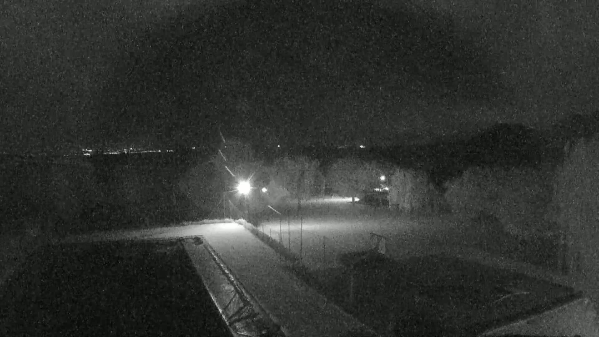 A nighttime, snowy scene shows a lightly snow-covered field with a few lights and what appears to be a vehicle in the distance.
