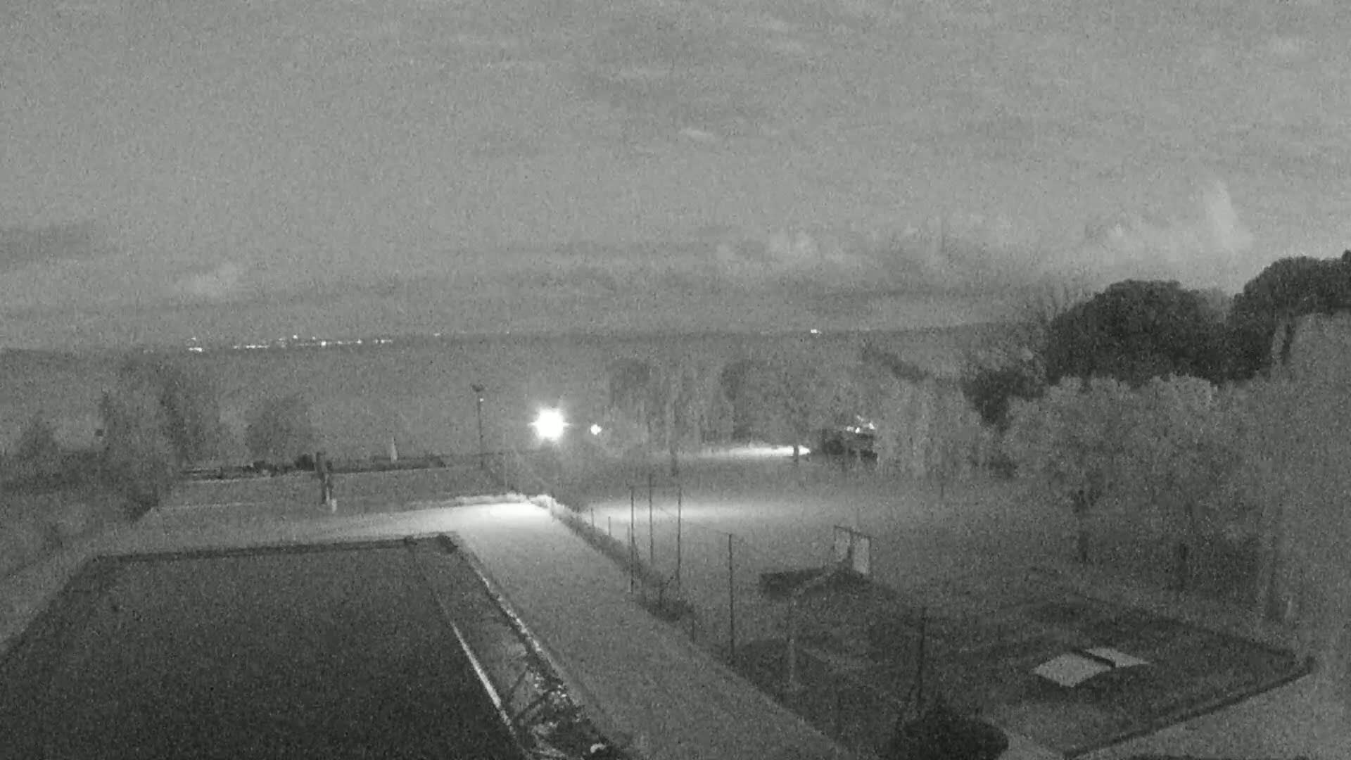 An outdoor nighttime scene shows an empty sports area with a pool and distant city lights under a cloudy sky.