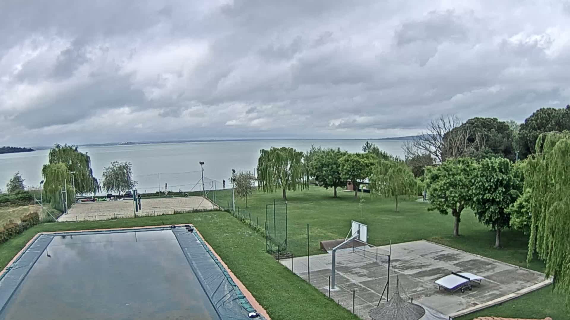 An overcast sky overlooks a grassy area with a swimming pool, volleyball court, basketball court, and ping pong table, all situated near a calm lake.