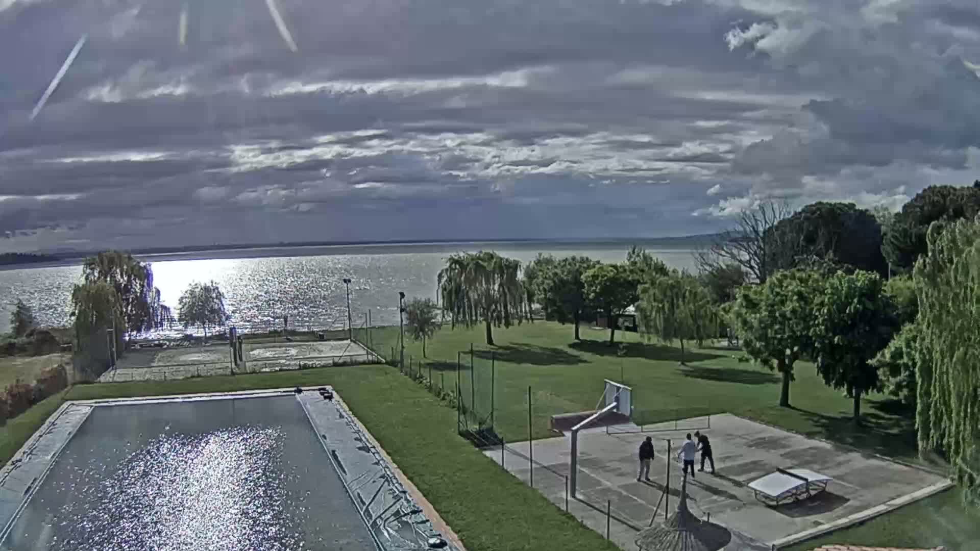 A swimming pool and sports courts are situated on a grassy area overlooking a calm lake under a partly cloudy sky.
