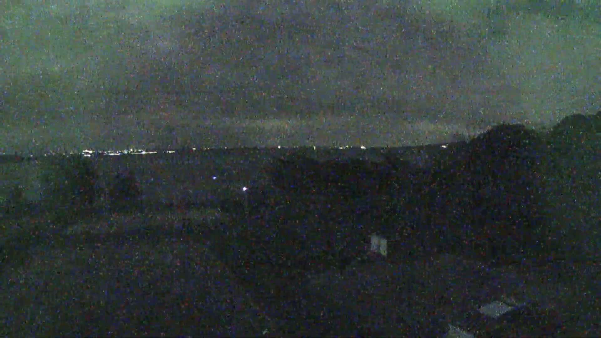 A nighttime landscape shows a dark, overcast sky above a hilly, dark terrain with distant city lights on the horizon.