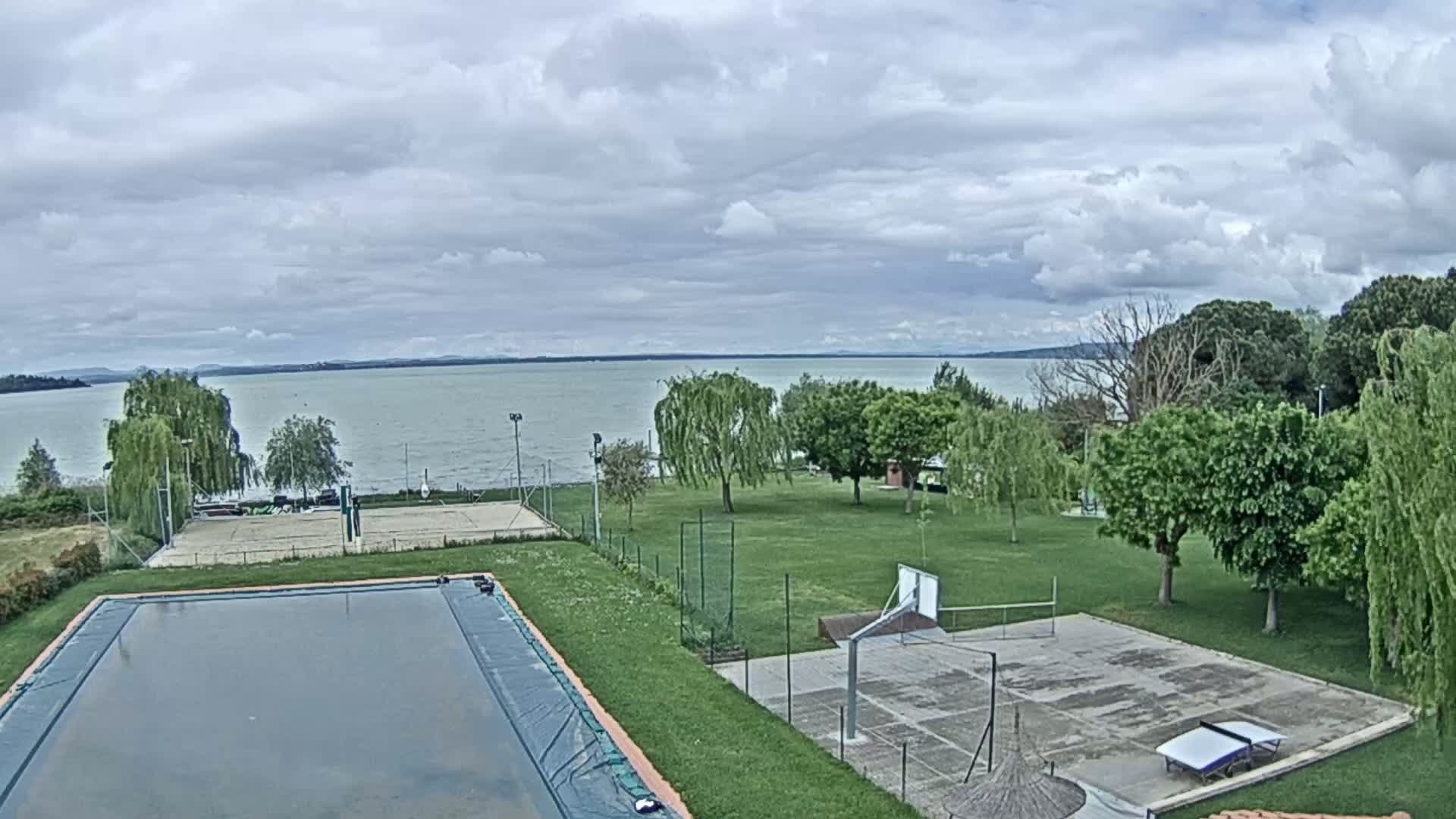 A swimming pool and sports courts sit beside a grassy area overlooking a calm lake under a cloudy sky.