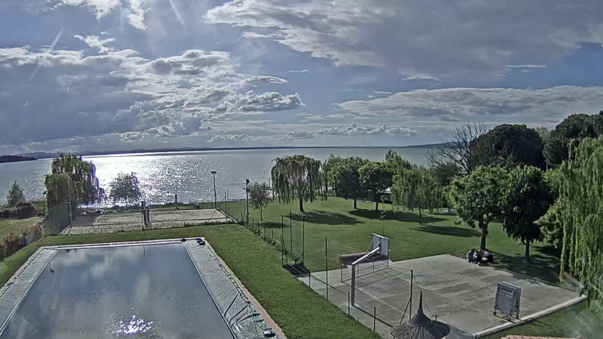 A sparkling lake is visible in the distance under a partly cloudy sky, with a swimming pool, grassy area, and a basketball court in the foreground.