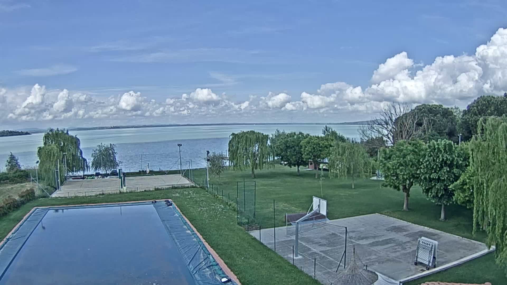 A swimming pool and outdoor courts sit on a grassy area overlooking a calm lake under a partly cloudy sky.