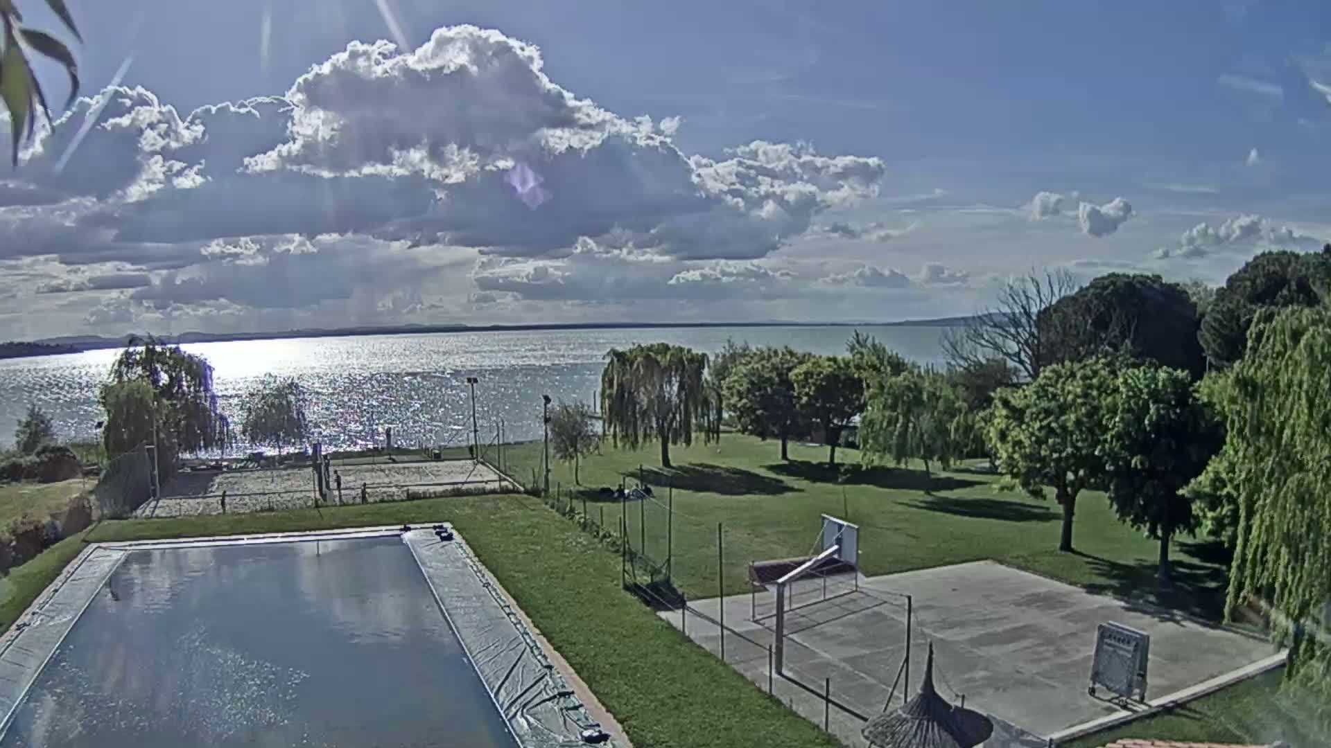 A swimming pool and sports courts are situated on a grassy area overlooking a sparkling lake under a partly cloudy sky.
