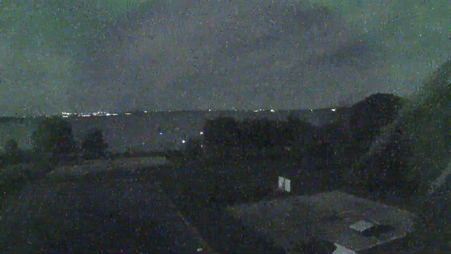 A nighttime view shows a dark, overcast sky over a line of distant city lights beyond a field of dark trees and a flat expanse of land.