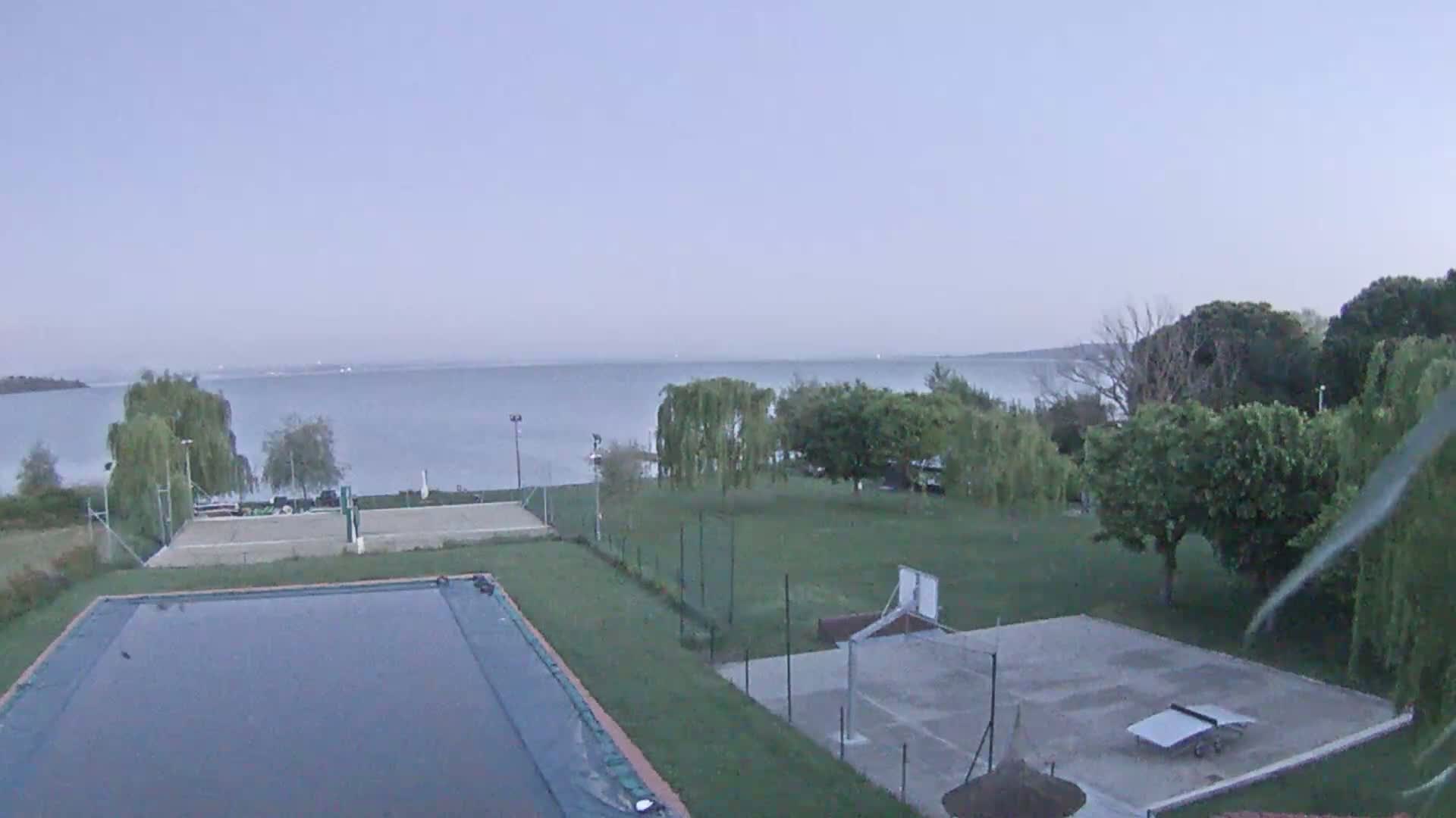 An outdoor scene shows a swimming pool, tennis courts, and a basketball court situated on a grassy area next to a calm lake under an overcast sky.