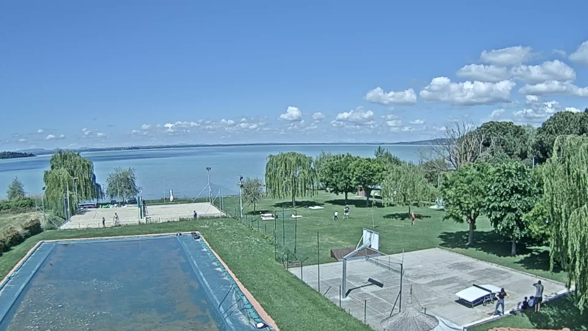 A swimming pool, volleyball court, basketball court, and ping-pong table are situated on a grassy area overlooking a calm lake under a partly cloudy sky.