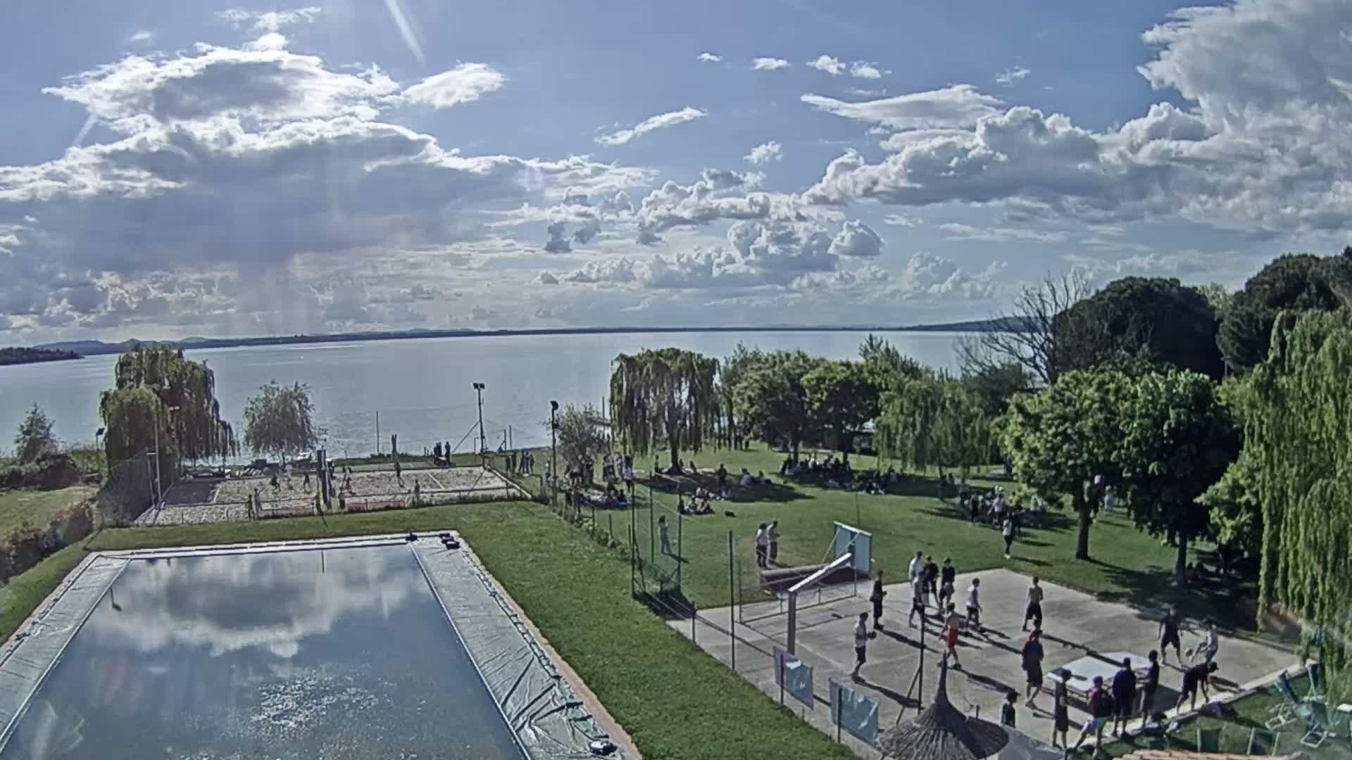 A partly sunny day shows a swimming pool, volleyball court, basketball court, and many people relaxing in a grassy area by a lake.