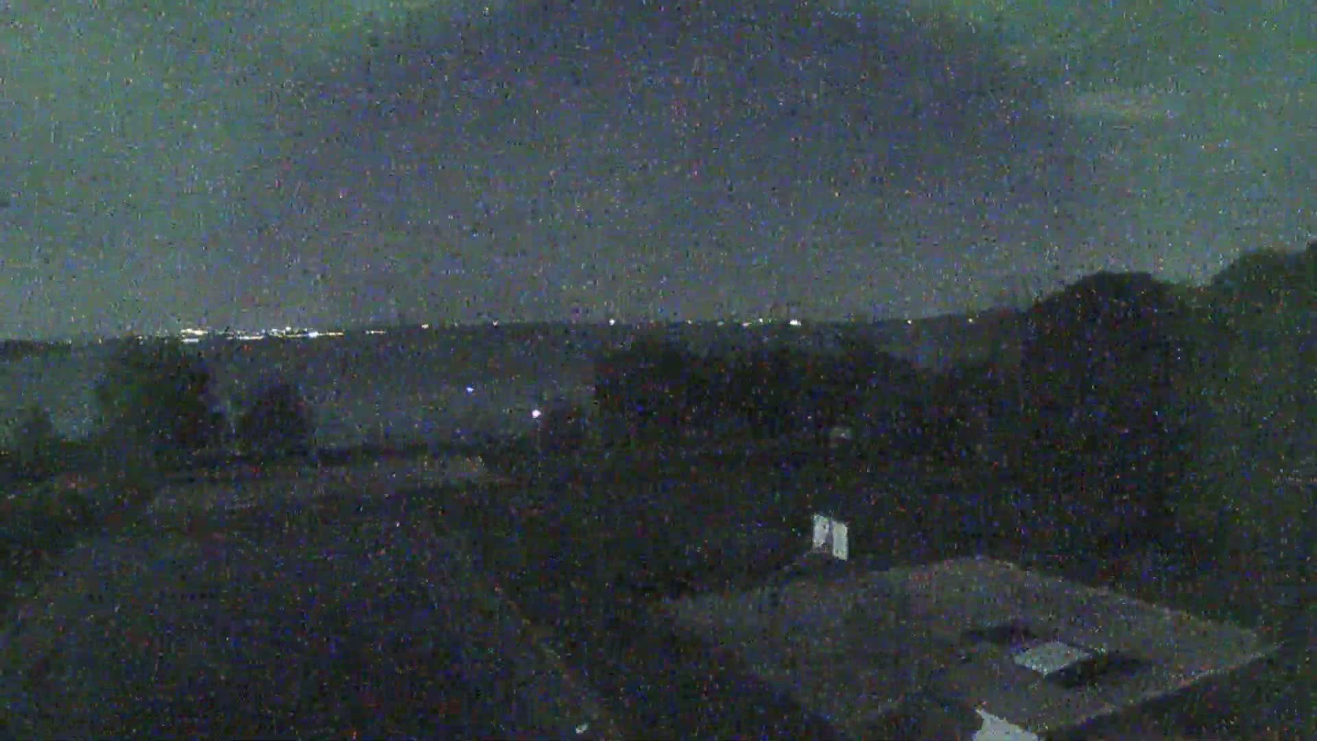 A nighttime view shows a dark landscape with distant city lights on the horizon under a cloudy sky.