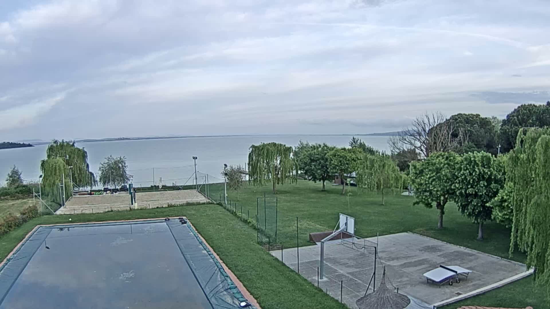 An outdoor scene shows a calm lake under a cloudy sky, with a grassy area containing a swimming pool, volleyball court, and basketball court.