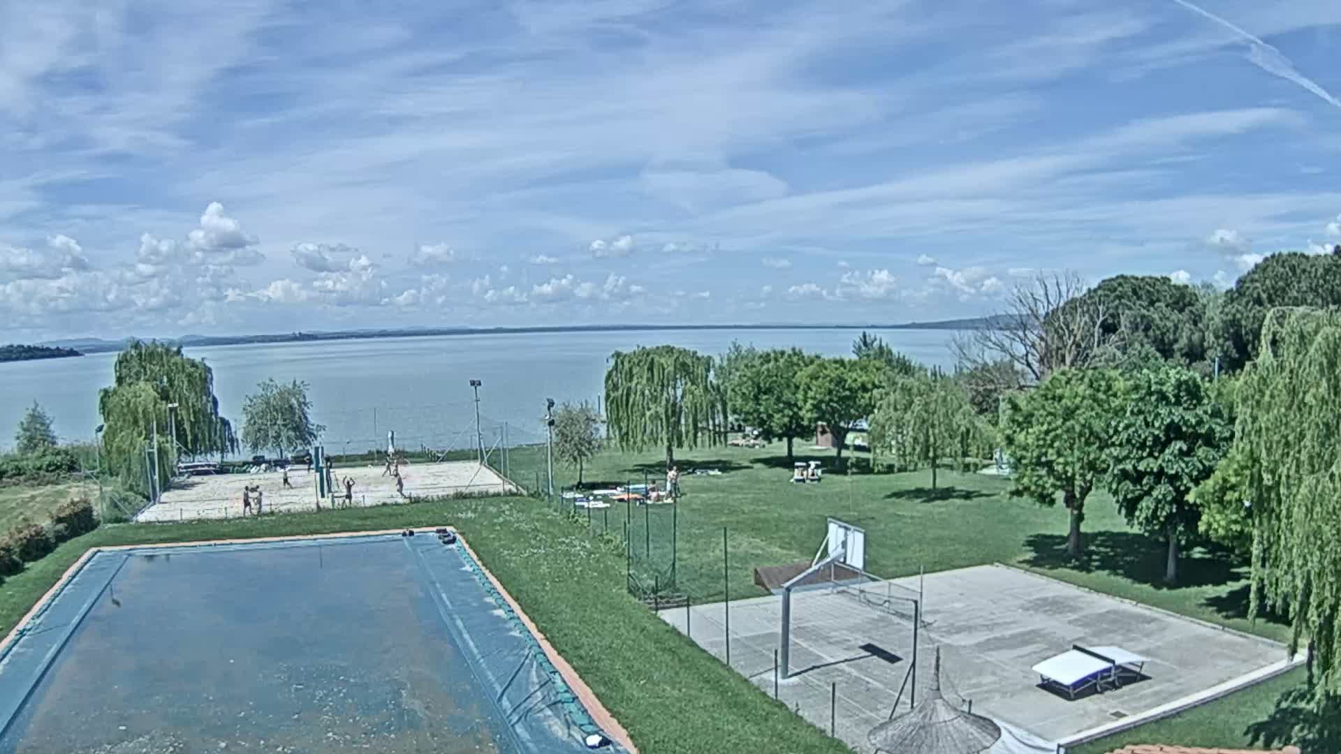 A swimming pool and various sports courts, including a volleyball court and a basketball court, are situated on a grassy area overlooking a calm lake under a partly cloudy sky.