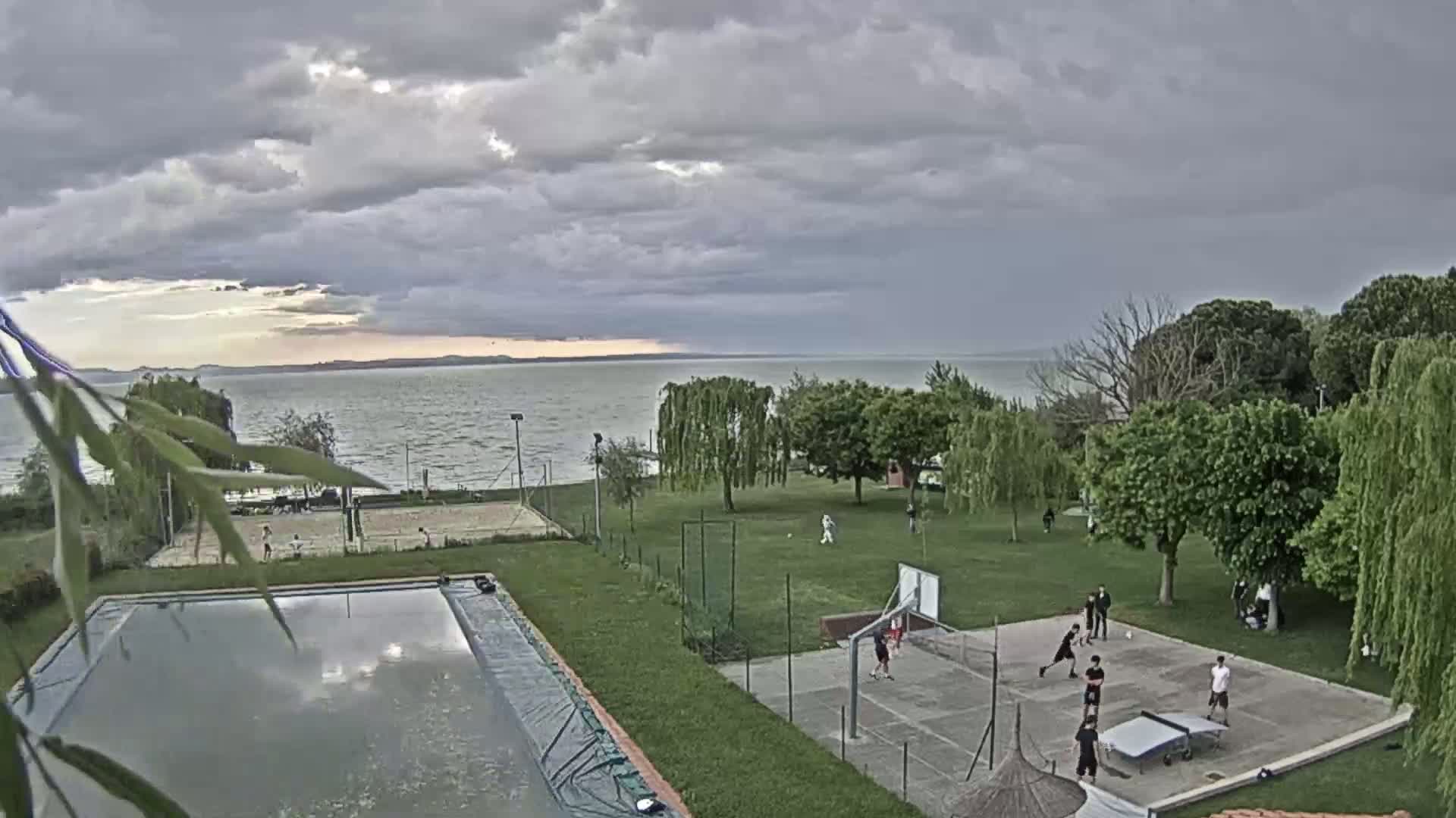 An outdoor scene shows a swimming pool, various sports courts with people playing, and a calm lake under a partly cloudy sky.