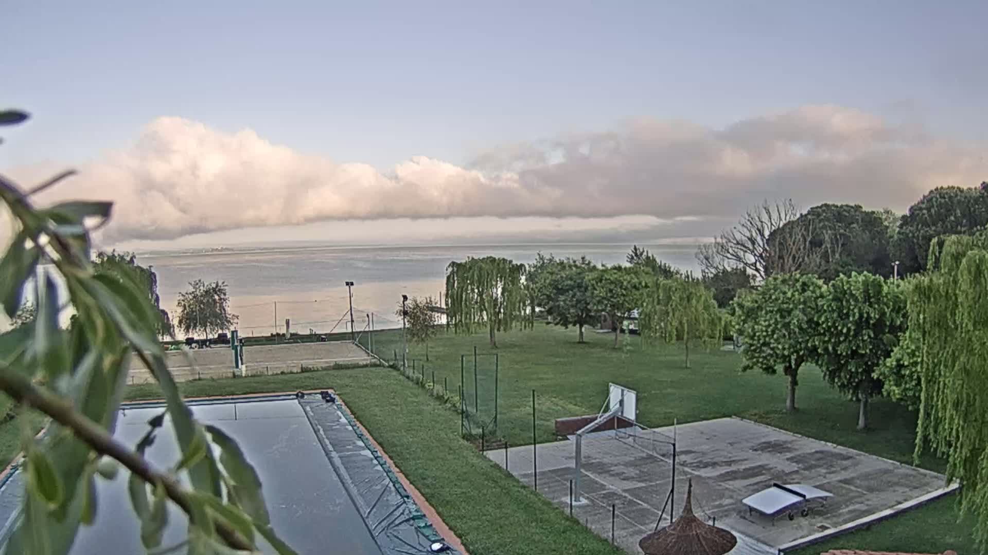 A calm lake is visible in the distance under a partly cloudy sky, behind a grassy area with a swimming pool, tennis court, and basketball court.
