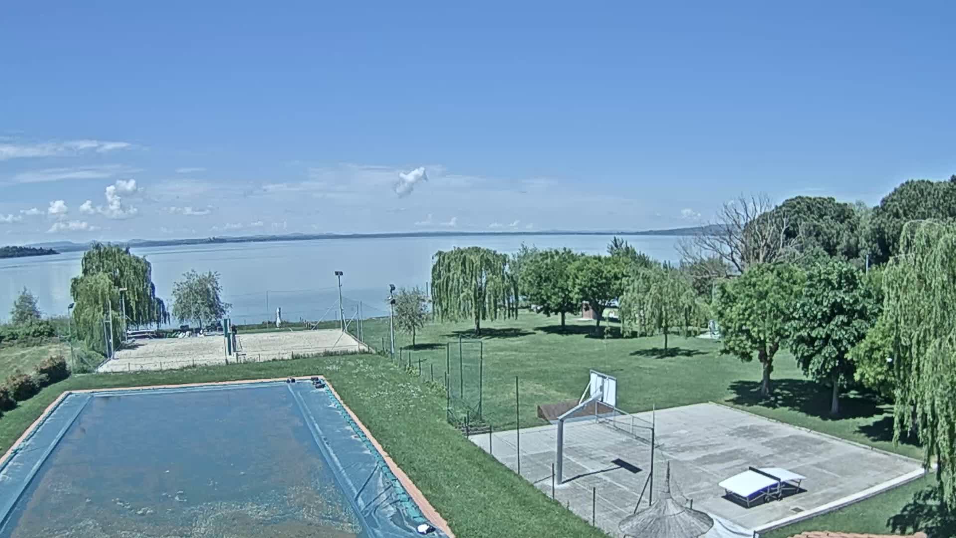 A swimming pool and outdoor courts for basketball and volleyball are situated on a grassy area next to a lake under a mostly sunny sky.