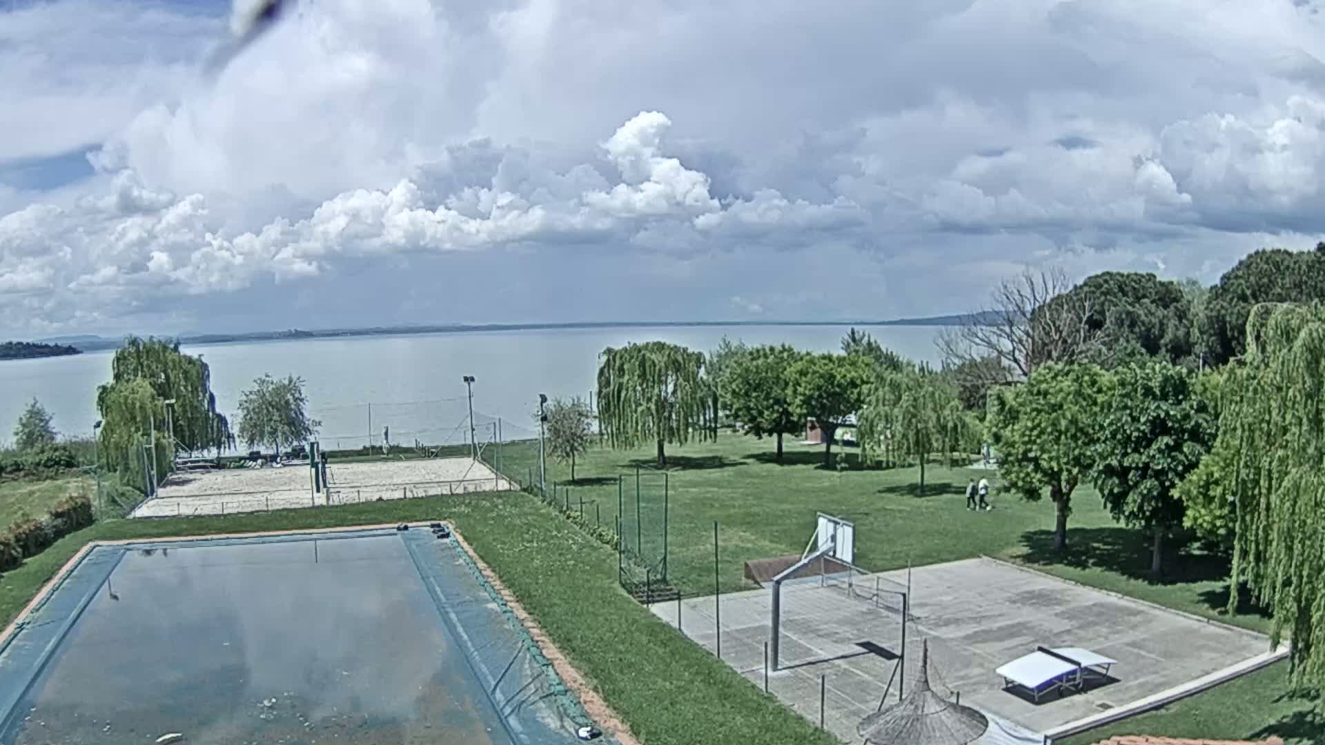 A swimming pool and outdoor courts for basketball and volleyball sit on a grassy area overlooking a calm lake under a partly cloudy sky.