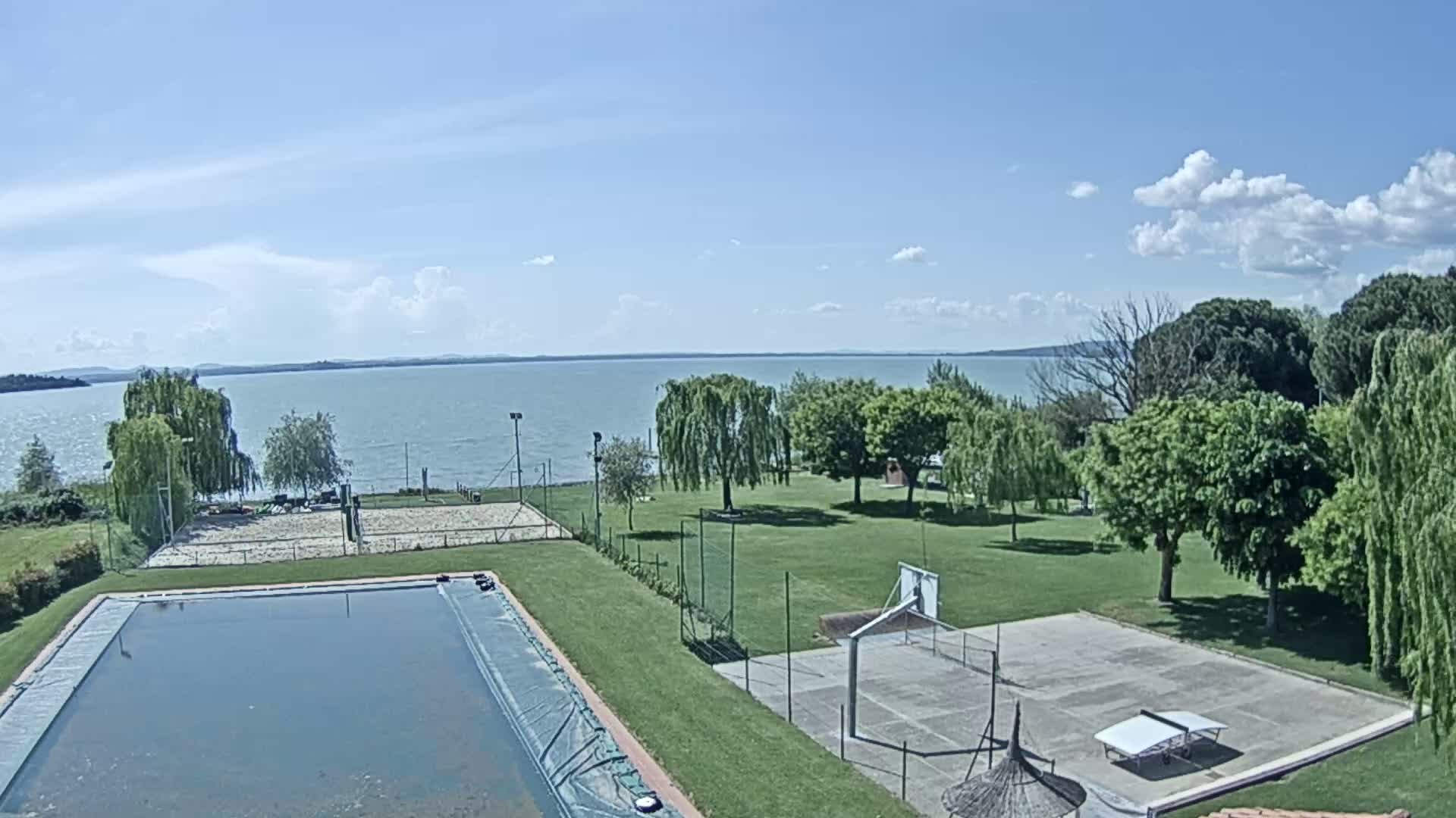 A swimming pool and several outdoor courts are situated on a grassy area next to a lake under a partly cloudy sky.