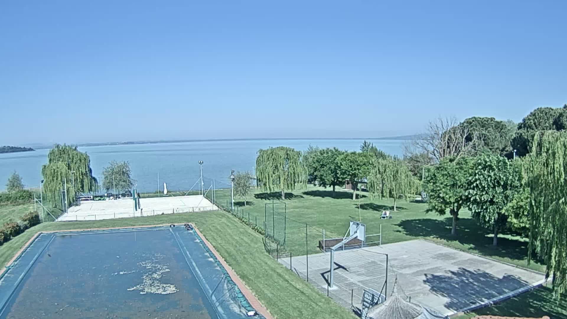 A clear, sunny day overlooking a calm lake with a volleyball court, basketball court, and empty swimming pool situated on a grassy area amongst trees.