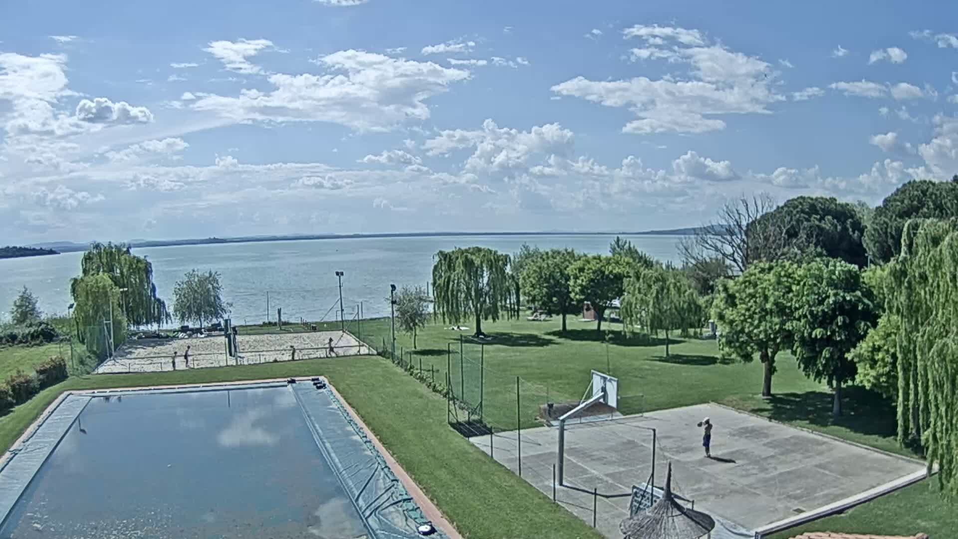 A swimming pool and outdoor courts for volleyball and basketball are situated on a grassy area overlooking a calm lake under a partly cloudy sky.