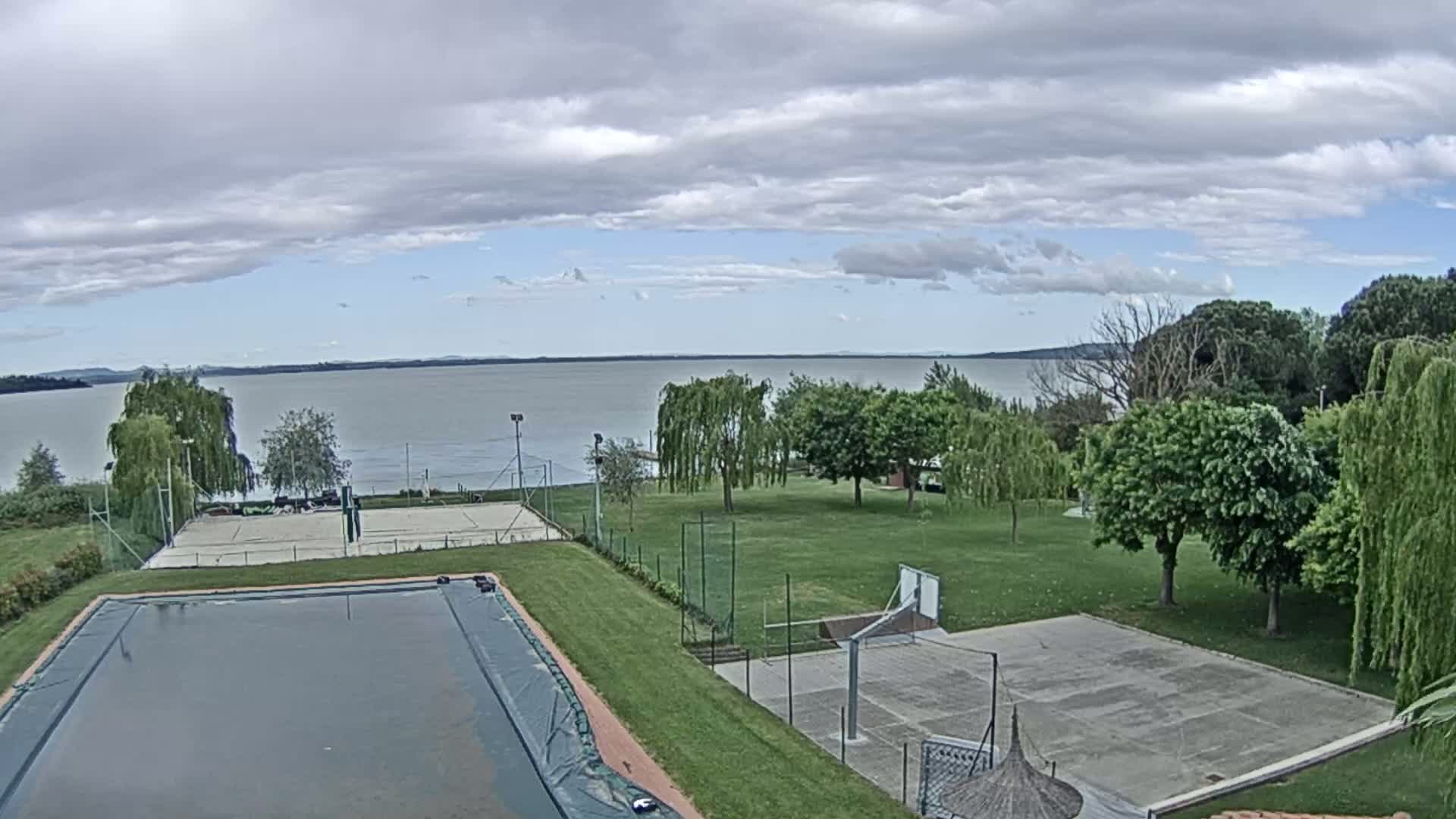 An outdoor view shows a covered swimming pool, tennis courts, and a basketball court situated on a grassy area overlooking a calm lake under a partly cloudy sky.