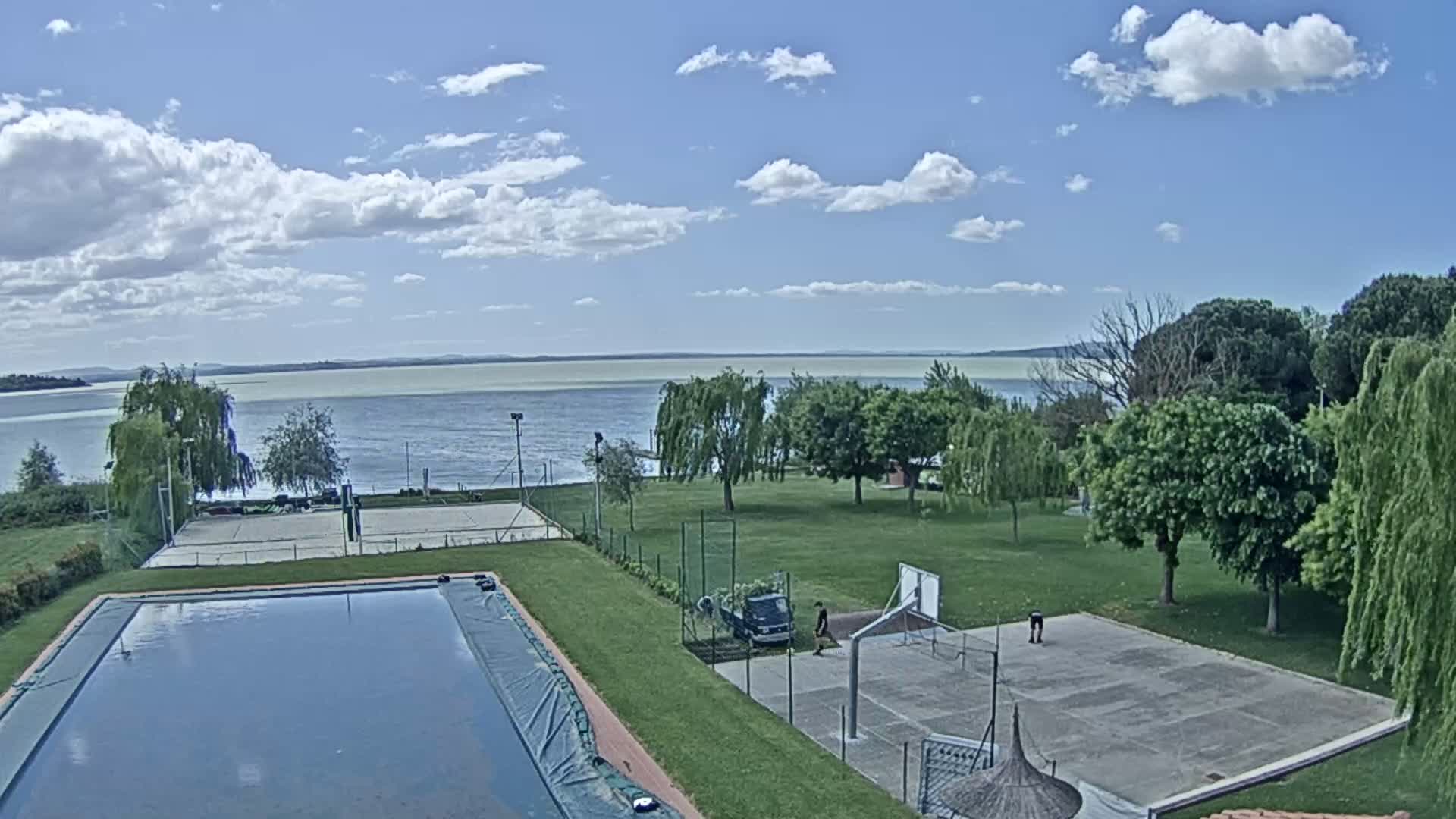 A swimming pool and sports courts are situated on a grassy area overlooking a calm lake under a partly cloudy sky.