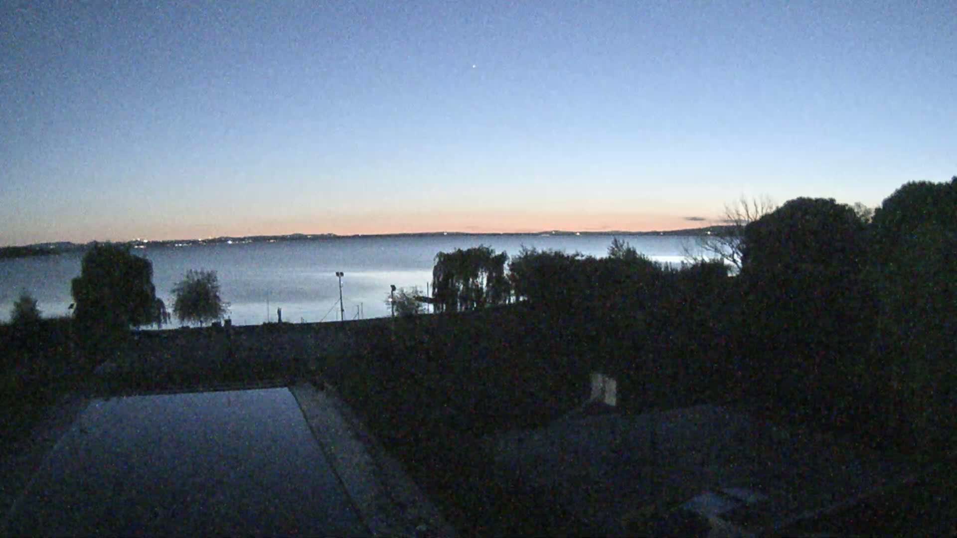 A calm lake at dusk is seen from a vantage point above, with dark trees and a partially visible pool in the foreground.