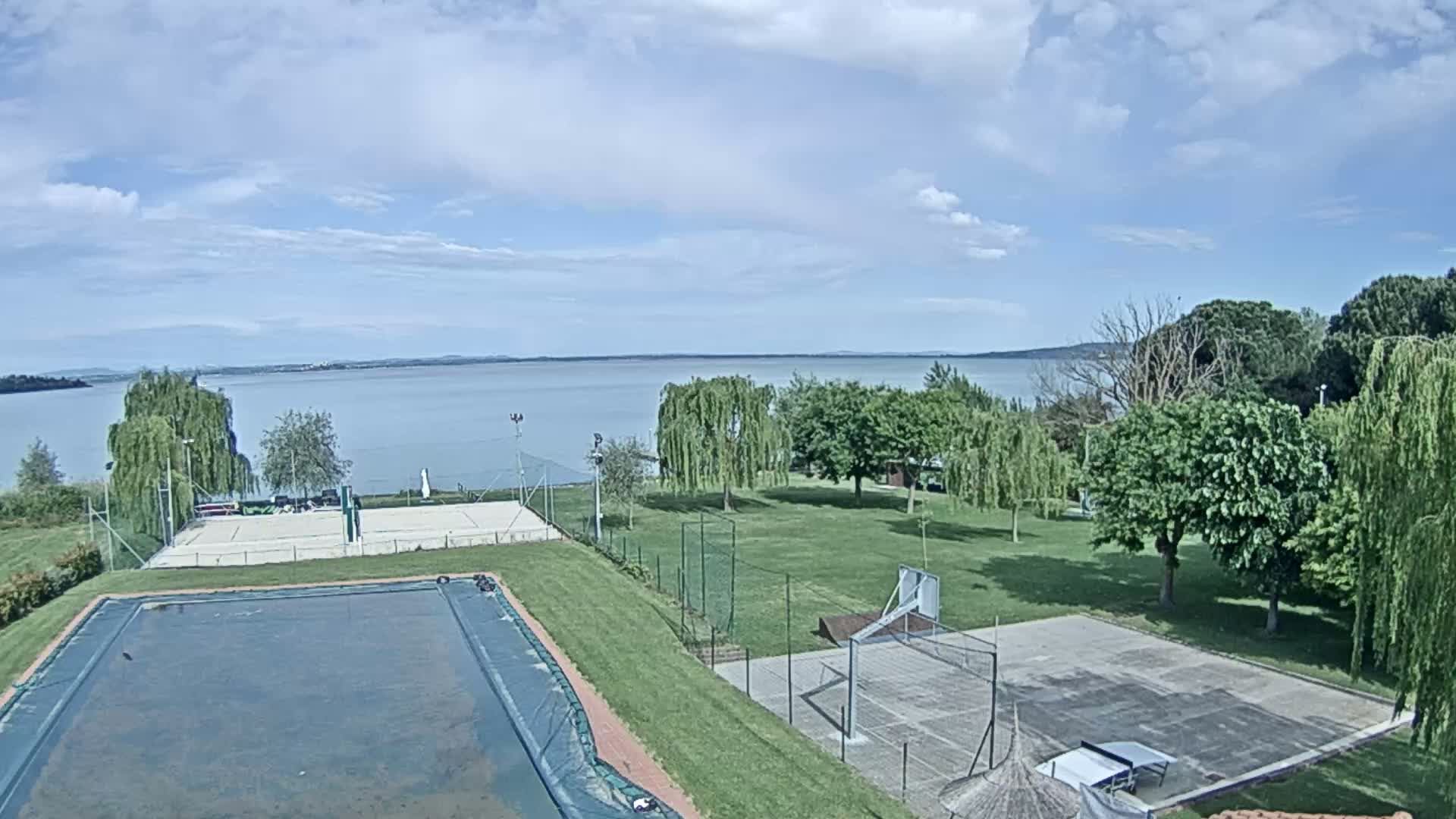 An outdoor scene shows a covered swimming pool, tennis and basketball courts, and a large grassy area overlooking a calm lake under a partly cloudy sky.