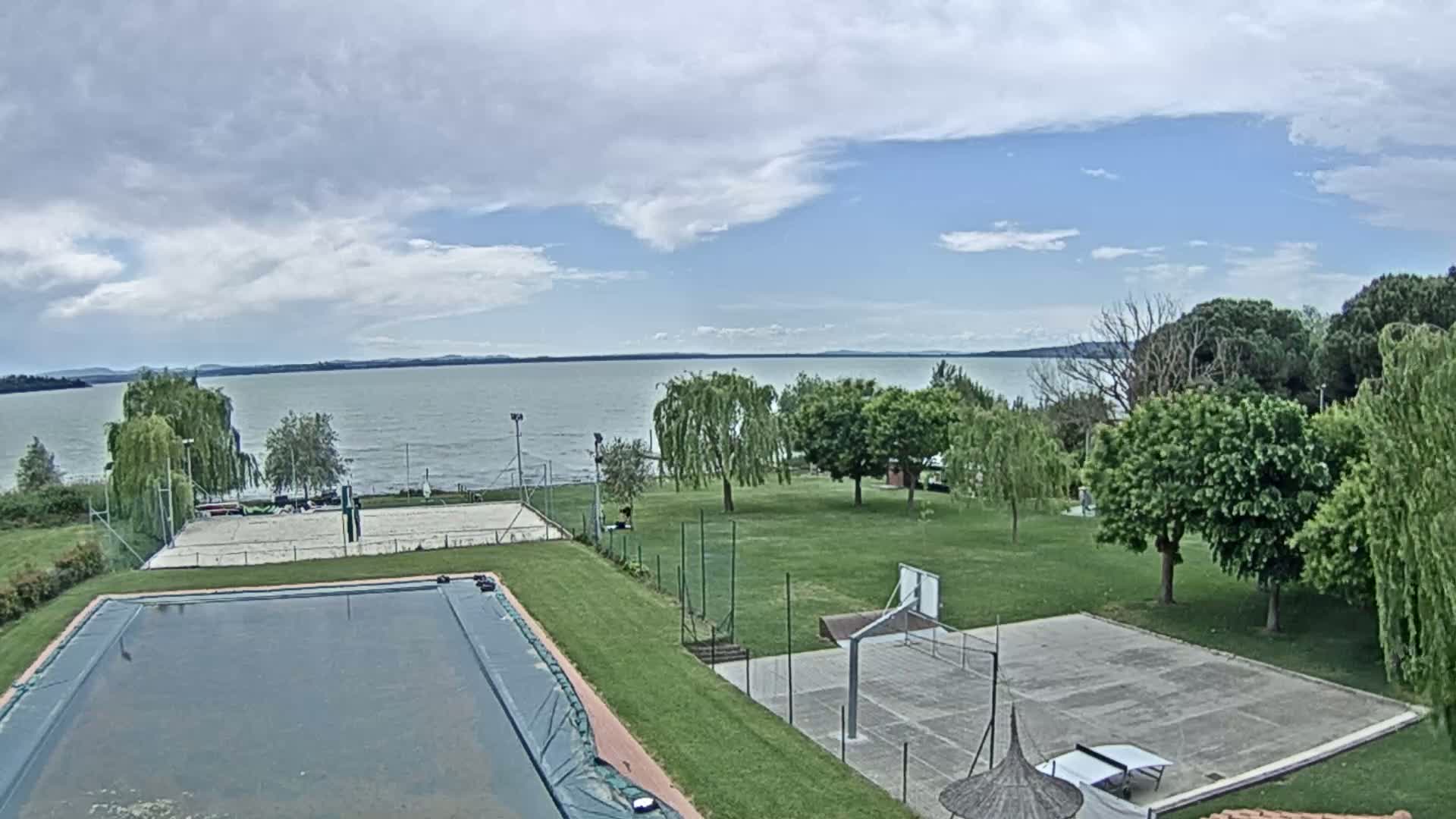 An outdoor view shows a calm lake under a partly cloudy sky, with a grassy area containing a pool, volleyball court, basketball court, and ping pong table.