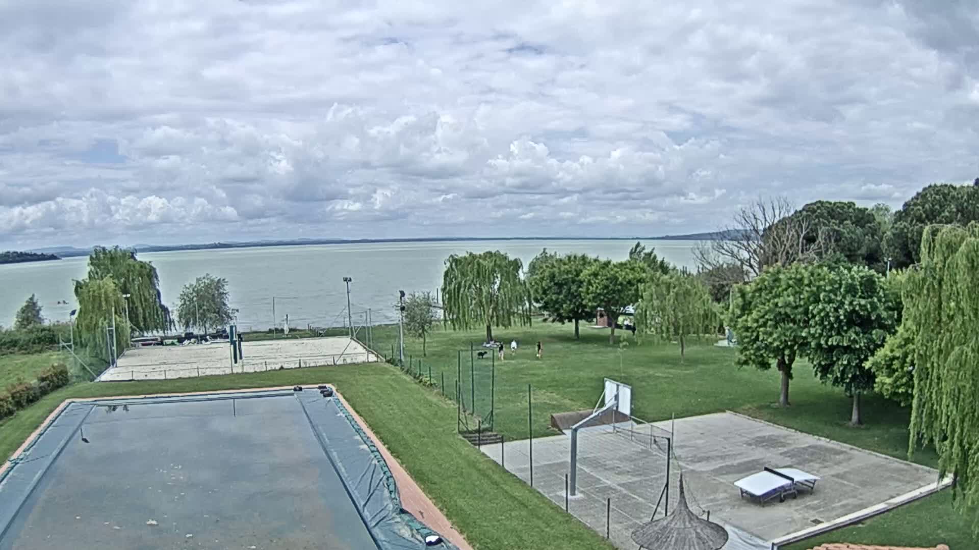 An outdoor scene shows a covered pool, volleyball court, basketball court, ping-pong table, and grassy area with trees, all overlooking a calm lake under a partly cloudy sky.