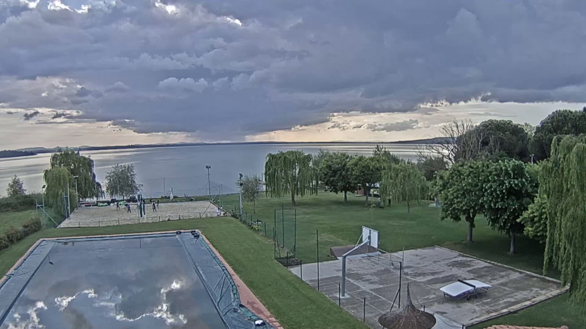 An overcast sky hangs over a lakeside property featuring a swimming pool, volleyball court, basketball court, and ping-pong table.