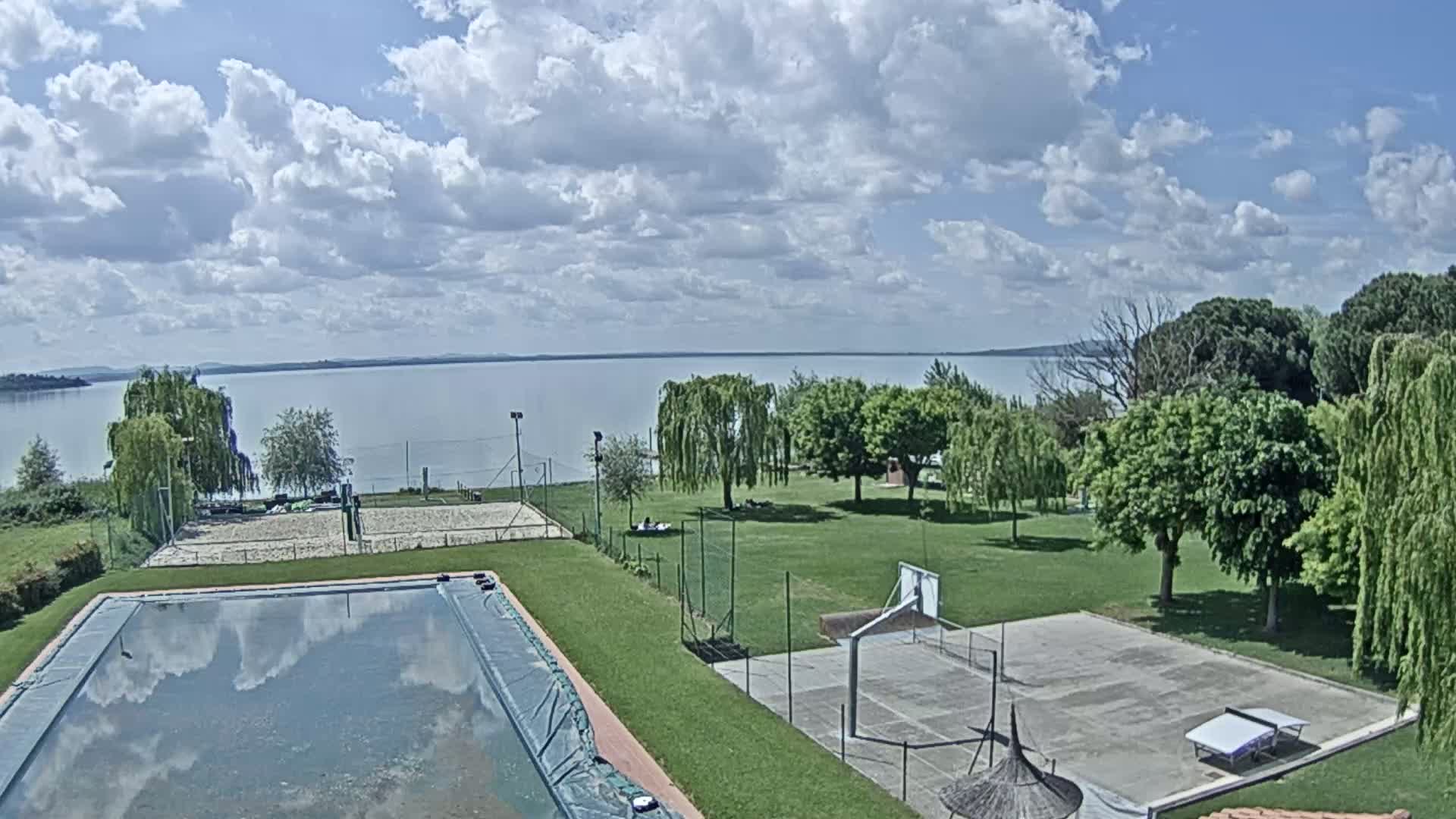 A swimming pool and outdoor courts sit on a grassy area overlooking a calm lake under a partly cloudy sky.
