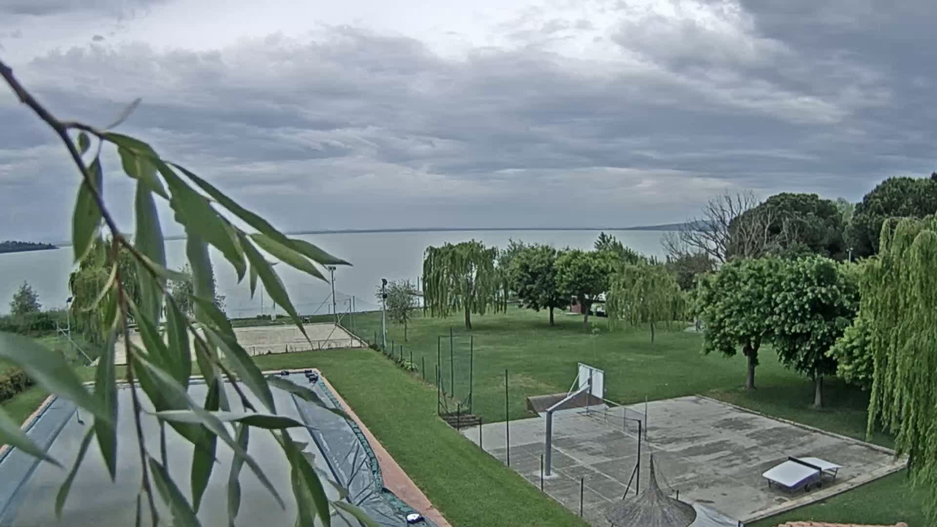 A covered swimming pool and a basketball court are situated on a grassy area overlooking a calm lake under a cloudy sky.