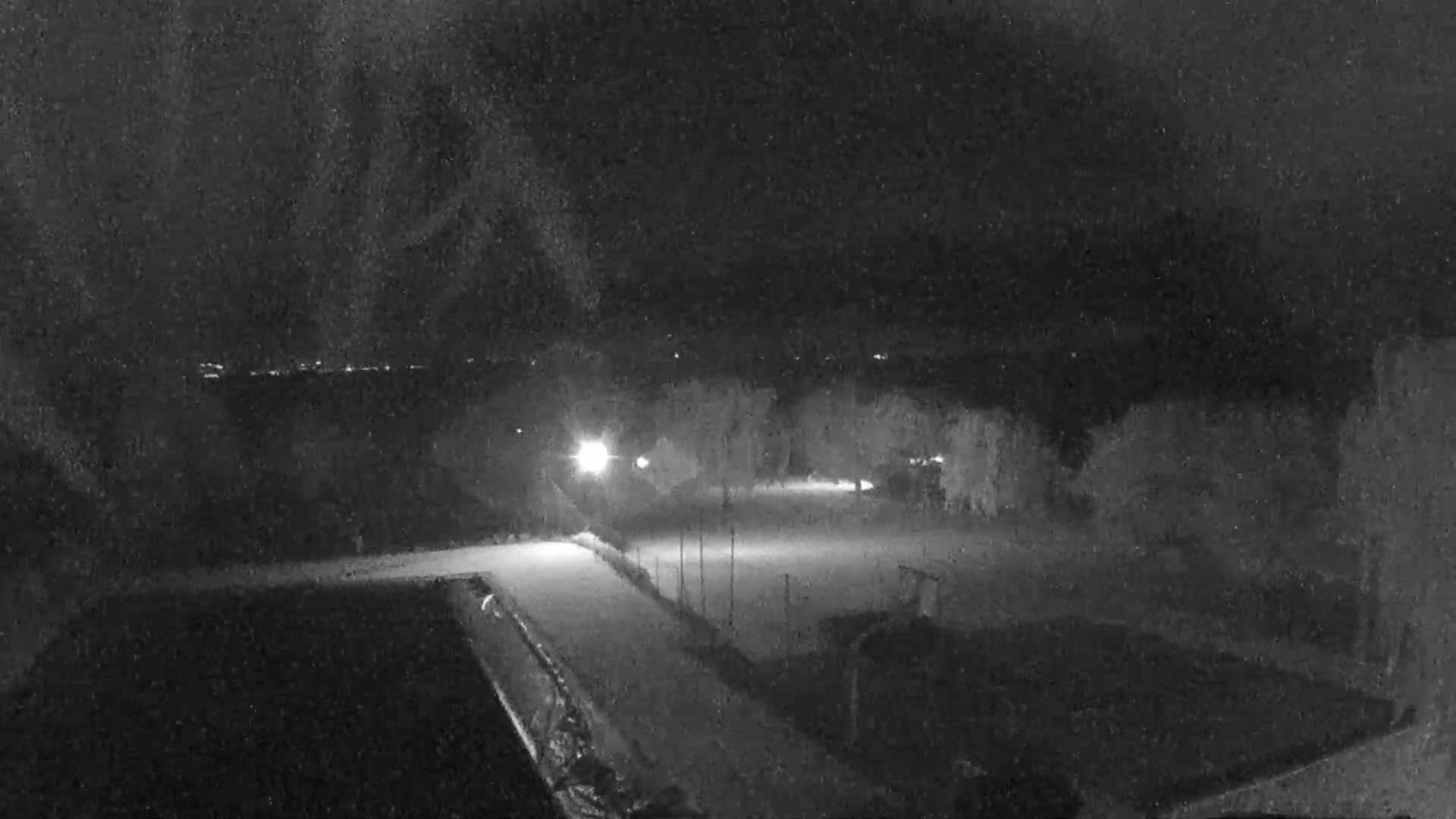A nighttime, snowy scene shows a brightly lit empty paved area, surrounded by dark trees and buildings under a dark, cloudy sky.