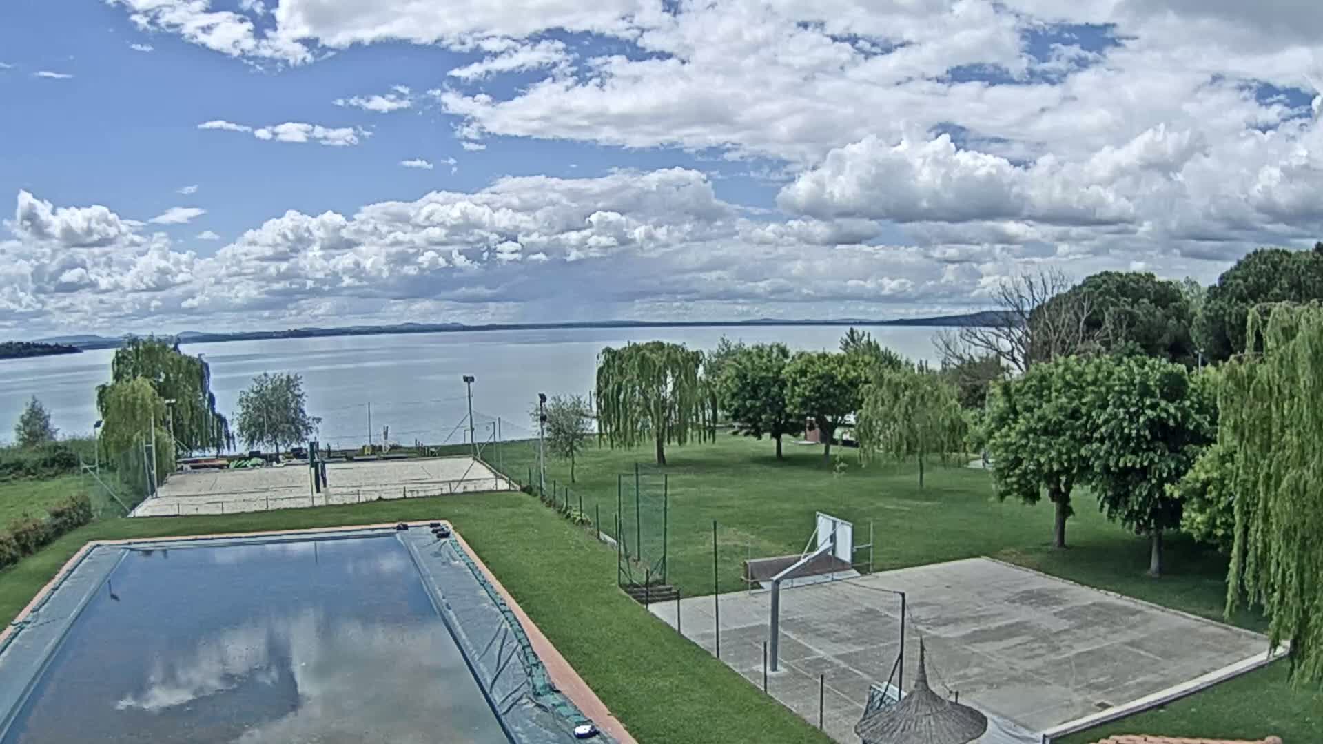 A swimming pool and outdoor sports courts are situated on a grassy area overlooking a calm lake under a partly cloudy sky.