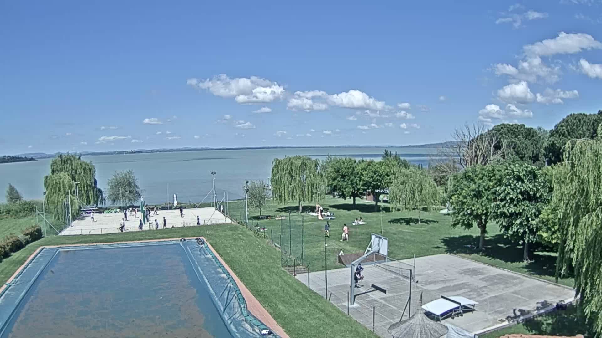 An outdoor scene on a partly sunny day shows a large body of water in the background, with a swimming pool, volleyball court, and basketball court in the foreground, where several people are playing sports and relaxing.