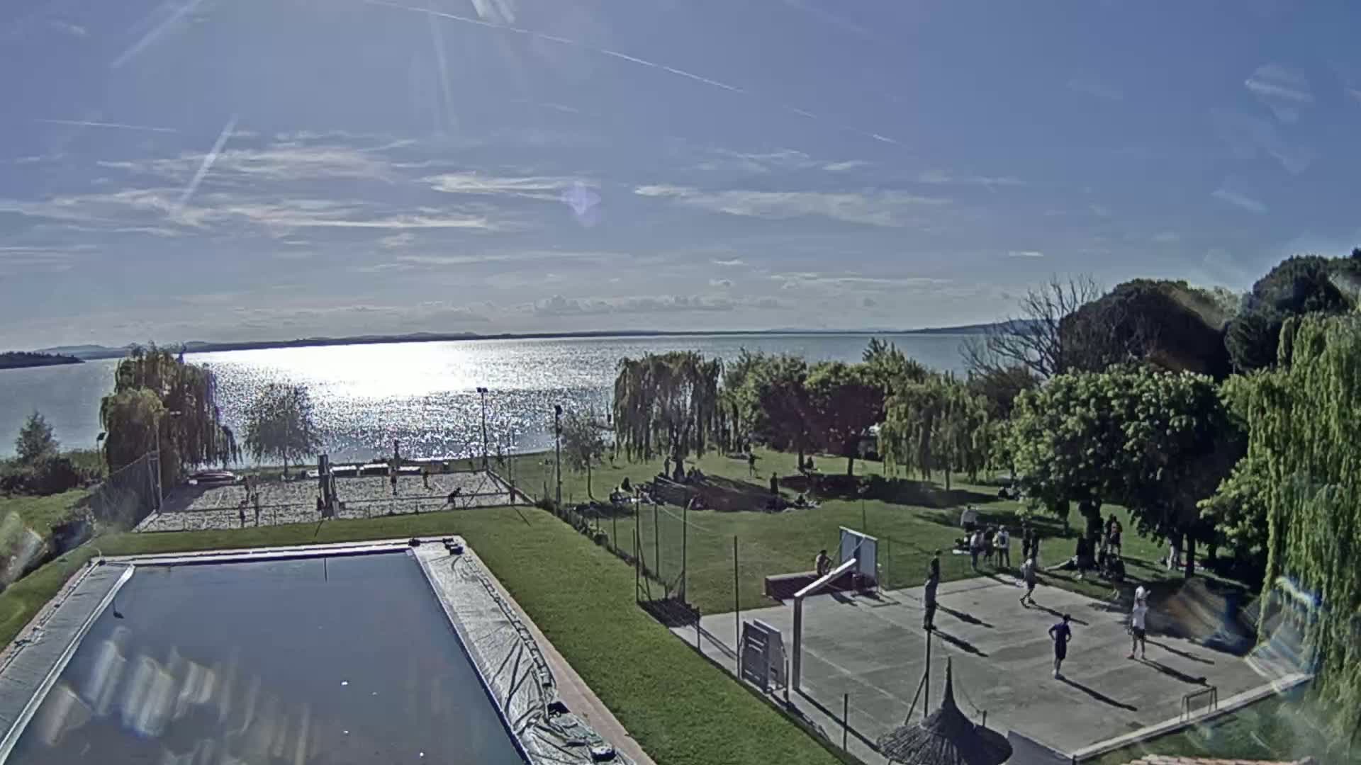 A sunny day overlooks a sparkling lake, with people enjoying recreational activities on the grassy shore including volleyball and basketball courts, near a swimming pool.