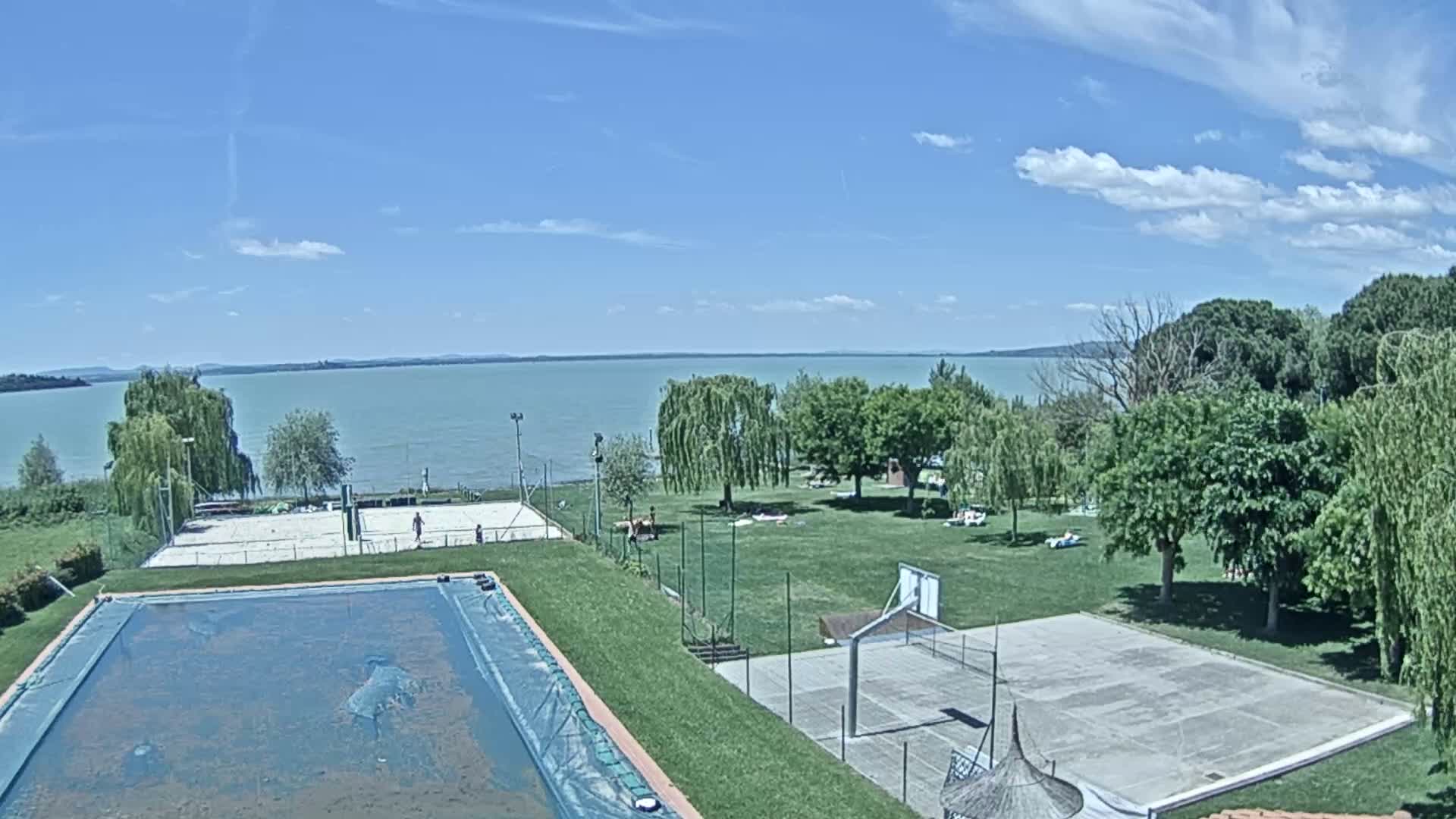 A swimming pool and sports courts are situated on a grassy area overlooking a calm lake under a partly cloudy blue sky.