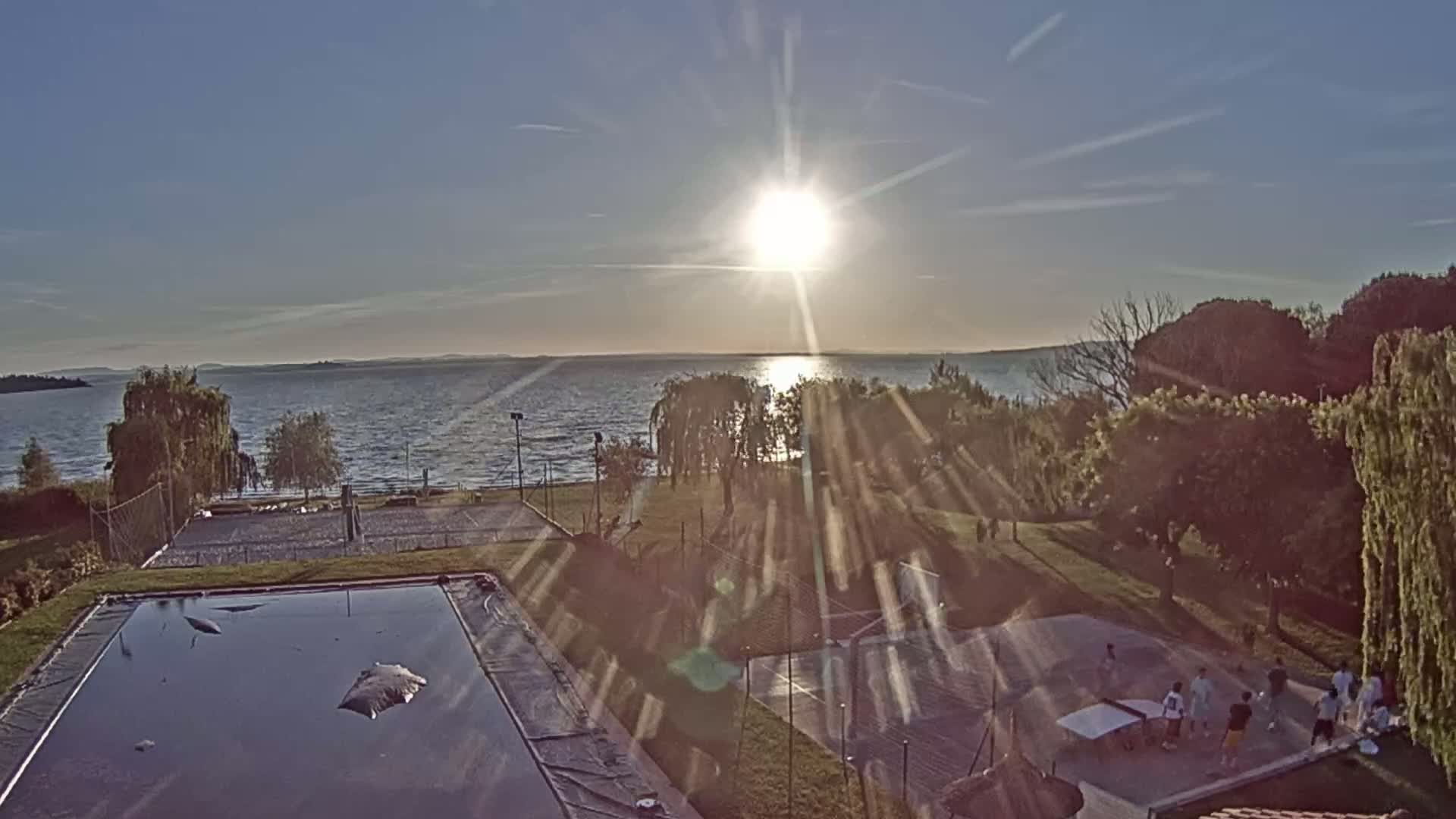 An aerial view shows a calm lake at sunset with a grassy area containing a swimming pool, tennis courts, and people playing ping pong.