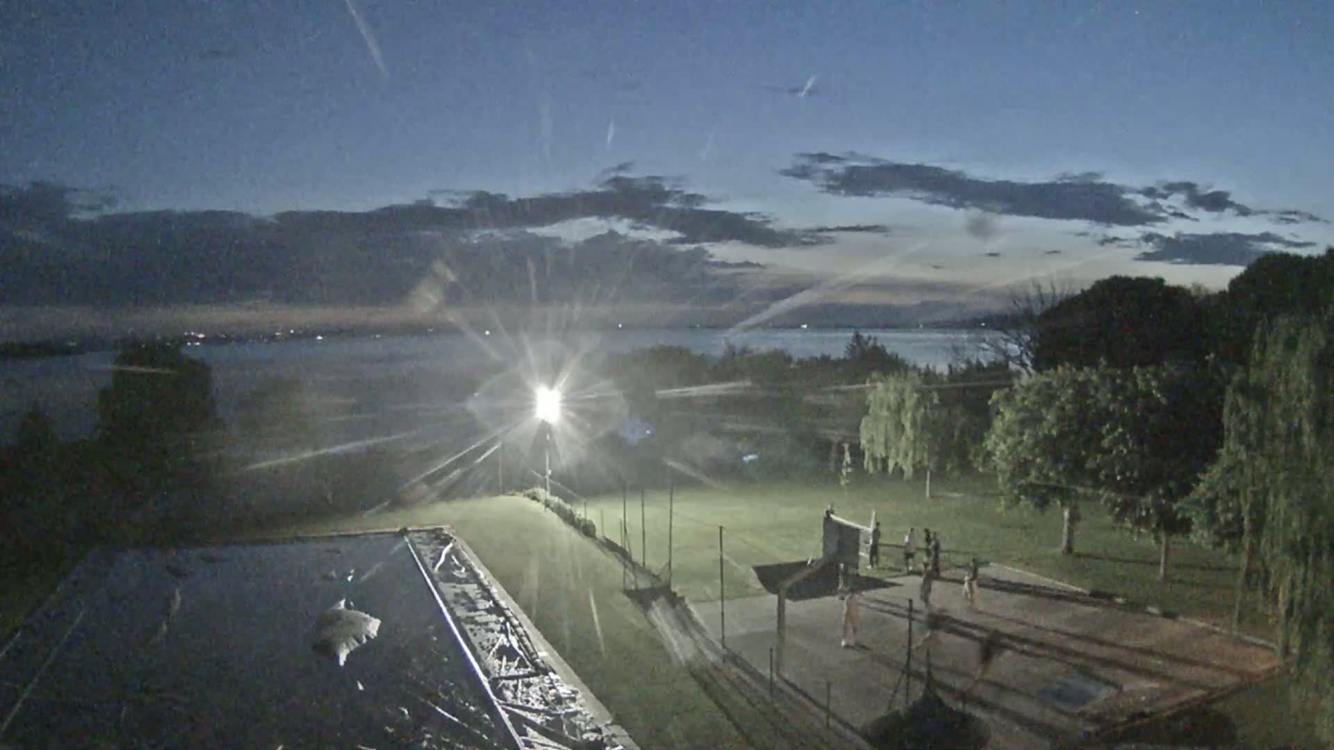 Under a twilight sky with scattered clouds, people play on a lighted outdoor court near a body of water.