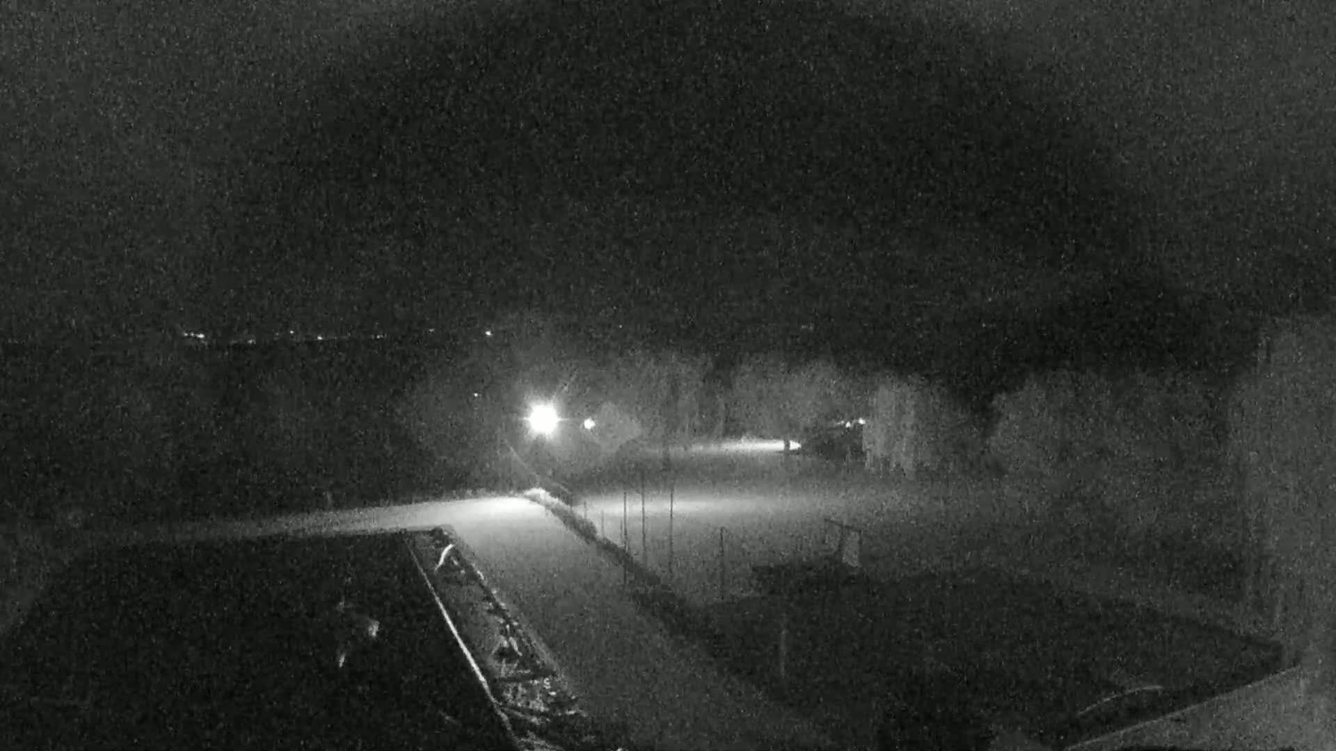 A nighttime, snow-covered area is illuminated by several lights, showing a flat, possibly paved surface, and some surrounding trees and structures.