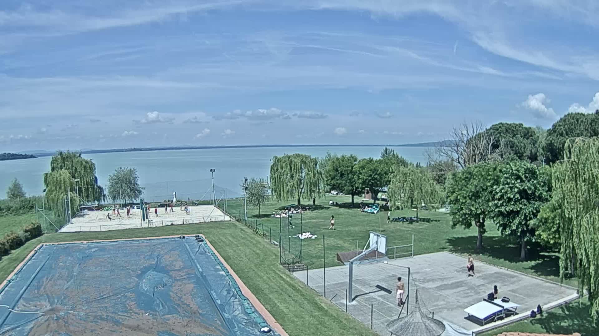 A sunny day overlooking a lake shows a volleyball court, basketball court, swimming pool, and people relaxing on a grassy area.