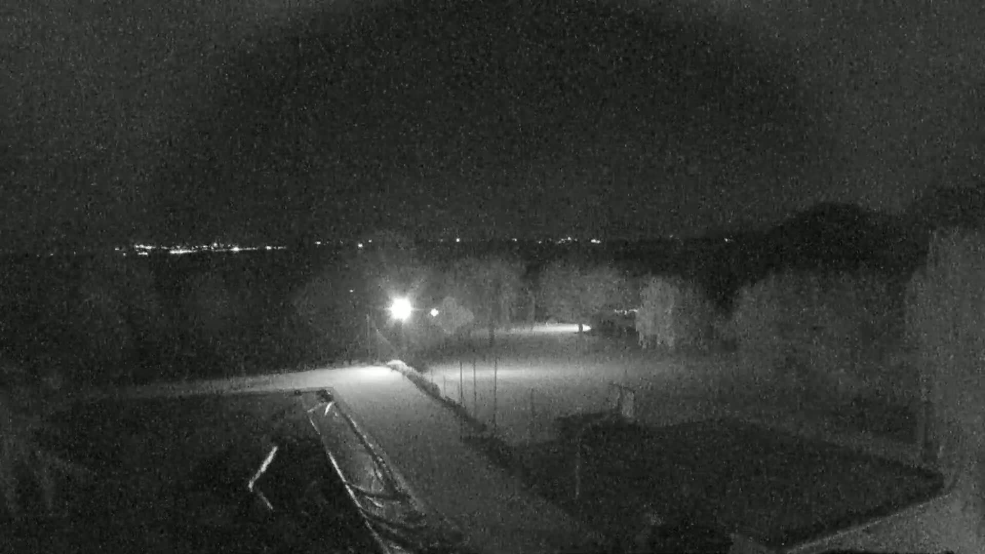 A nighttime, snowy scene shows a dimly lit area with some trees and buildings, a distant city skyline, and a possible sports field.
