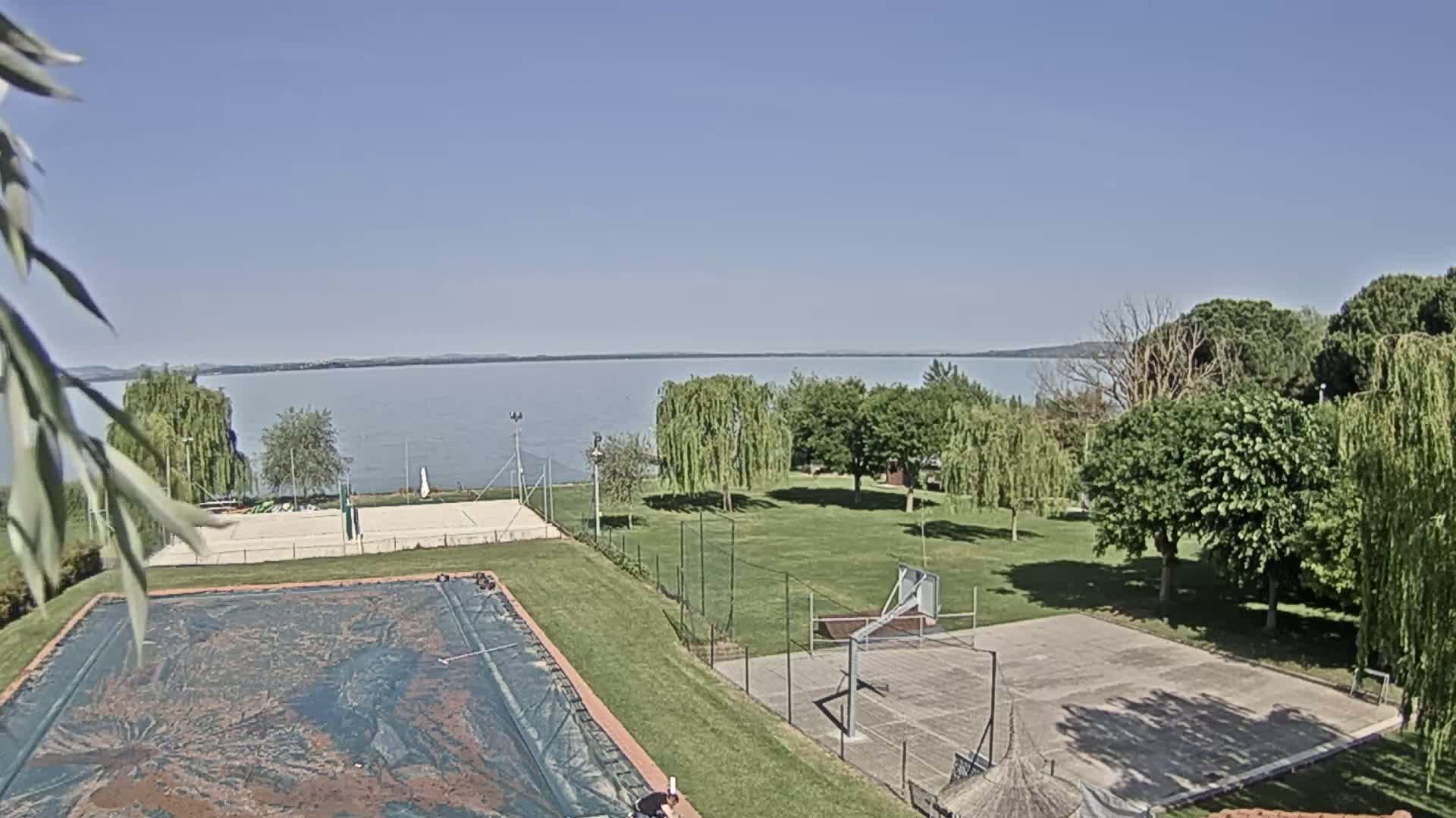 A clear, sunny day reveals a grassy area with a volleyball court, a basketball court, and a covered swimming pool, all overlooking a calm lake.