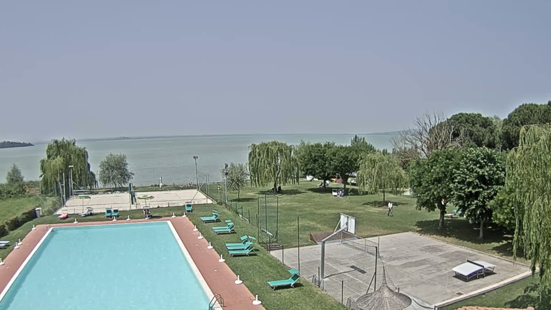 A swimming pool and recreational area with a basketball court and volleyball court, surrounded by trees and overlooking a calm lake under a clear sky.