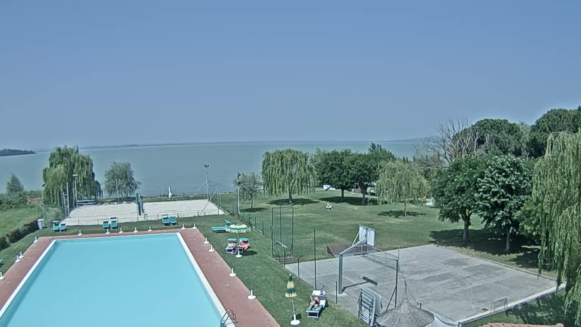 A swimming pool and outdoor courts sit on a grassy area next to a lake on a sunny day.