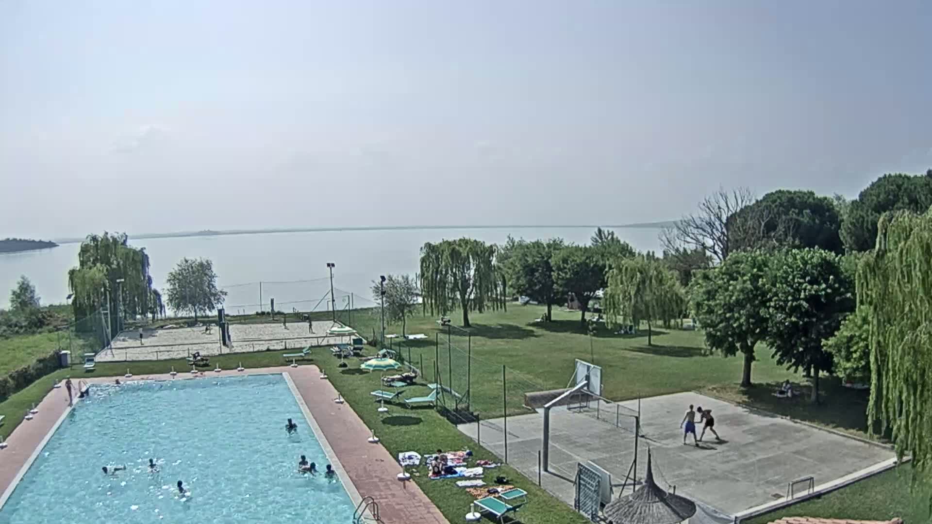 An outdoor swimming pool with people swimming in it is situated next to a volleyball court, a basketball court, and a grassy area overlooking a calm lake under a clear sky.