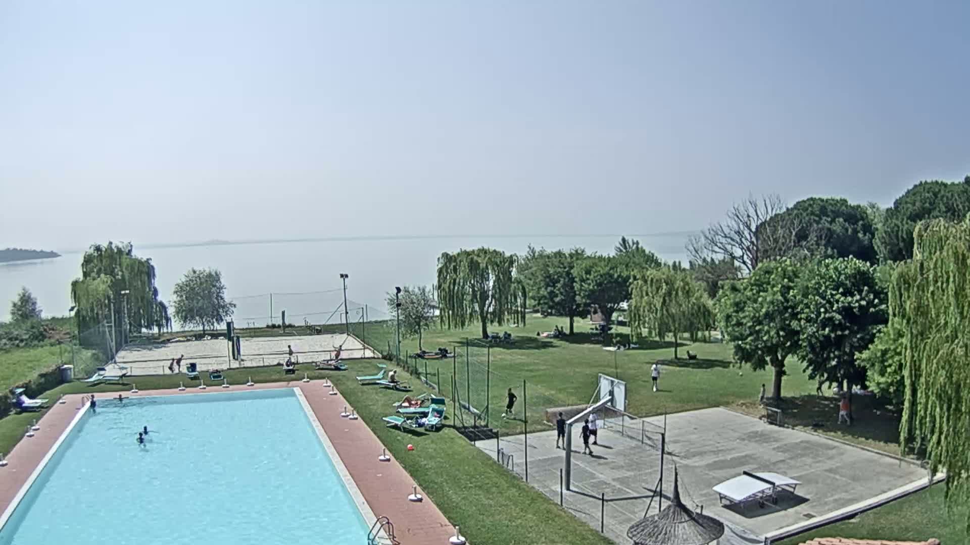 A swimming pool and recreational area with a volleyball court, basketball court, and ping pong table are situated on a grassy area overlooking a calm lake under a clear sky.