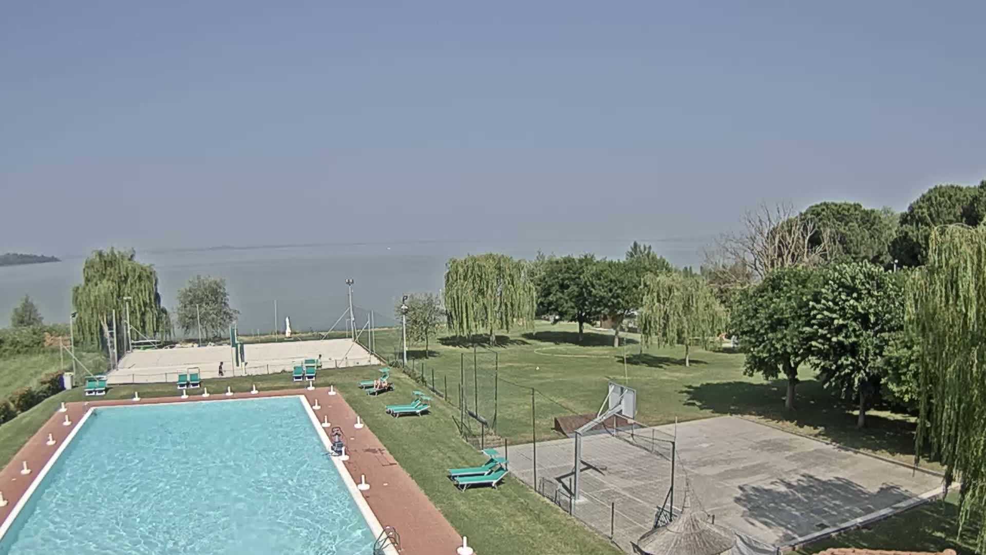 A swimming pool and sports courts are situated on a grassy area overlooking a calm lake under a clear sky.